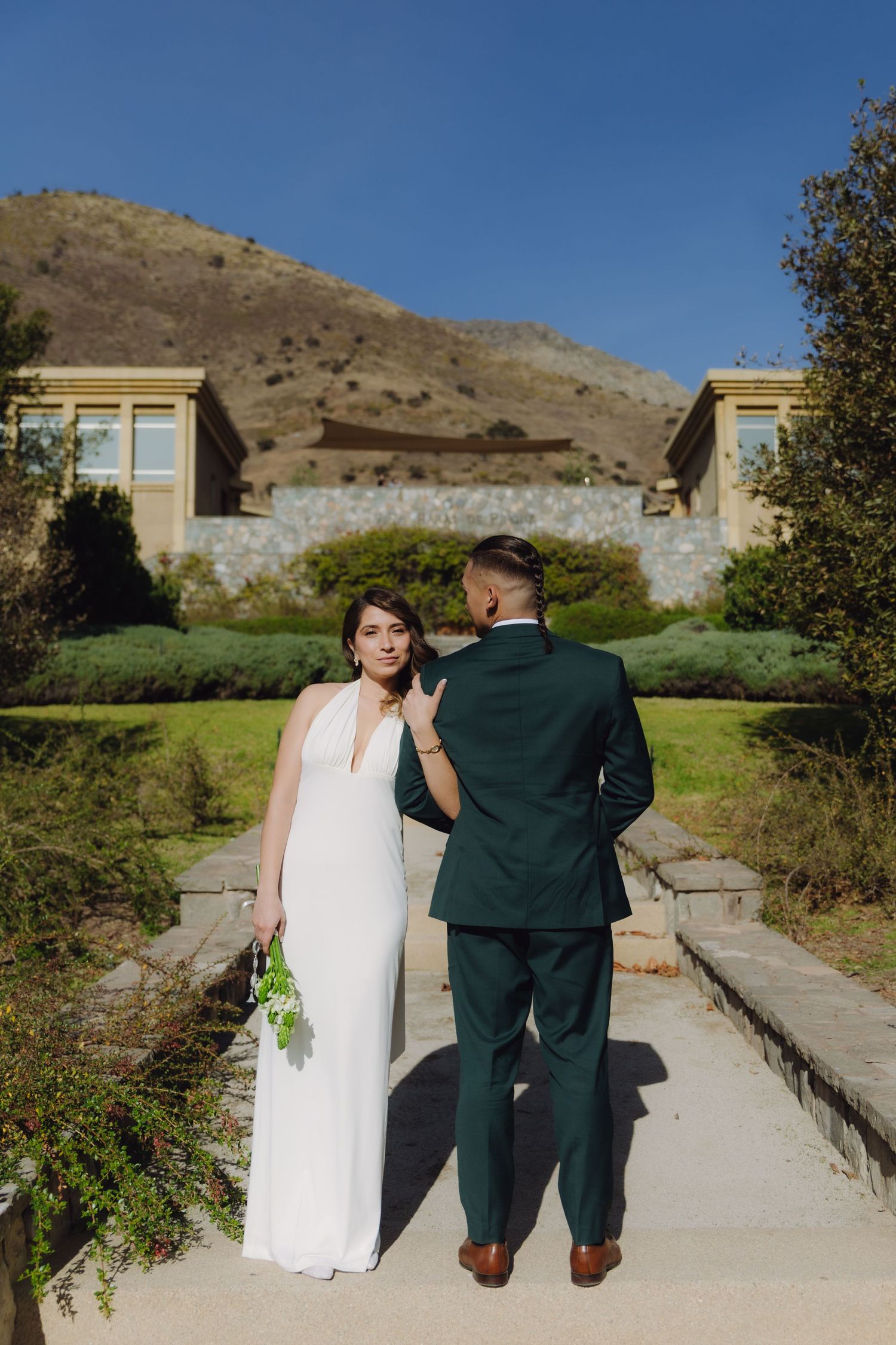 Bride in white halter gown looks back while groom in dark green suit stands facing a mountain estate venue.