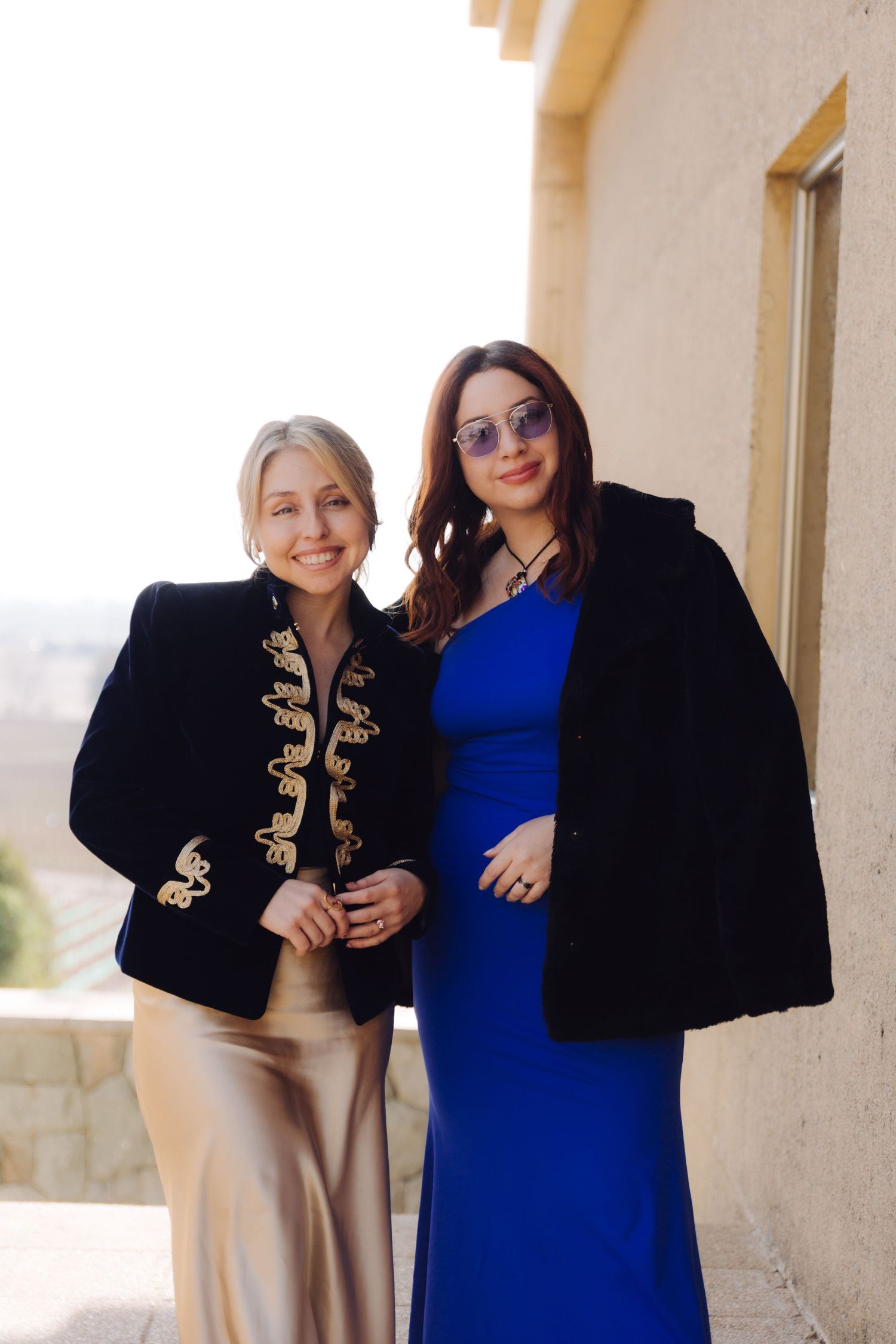 Two elegantly dressed women pose together outdoors, one in a gold skirt with embroidered jacket, other in a blue dress.