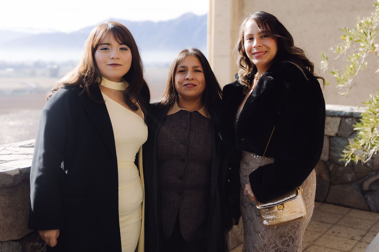 Three women pose together outdoors at a wedding venue with scenic mountain views in the background.