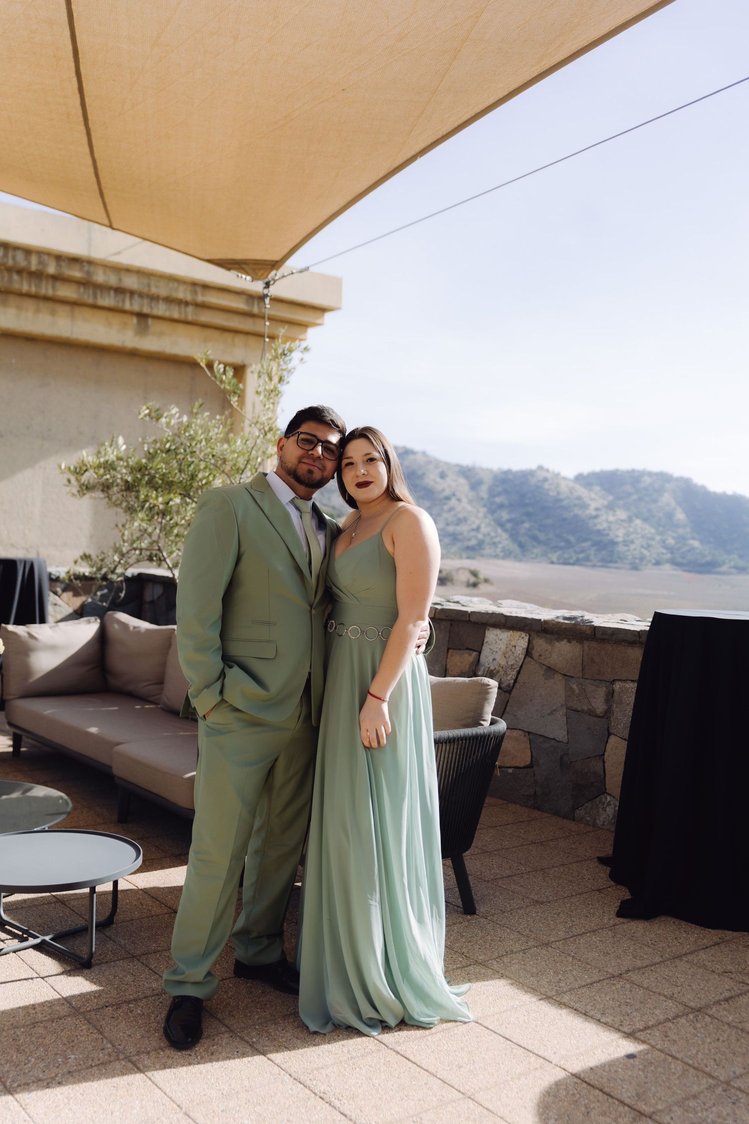 Couple in matching sage green formal attire posing on outdoor terrace with mountain views.