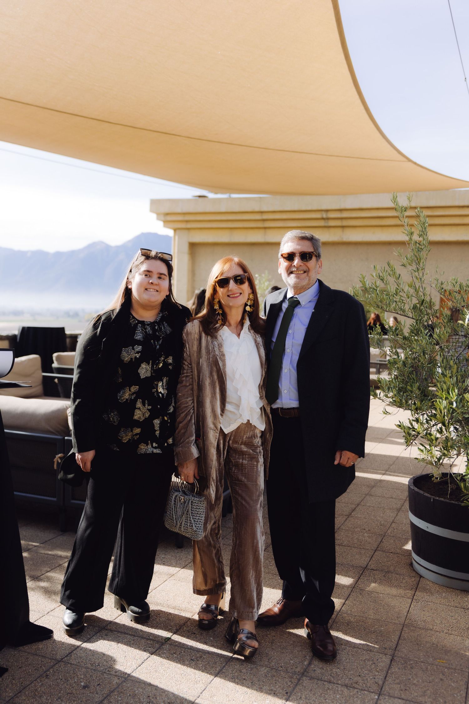 Three guests in stylish outfits posing together on sunny outdoor event terrace with mountains.