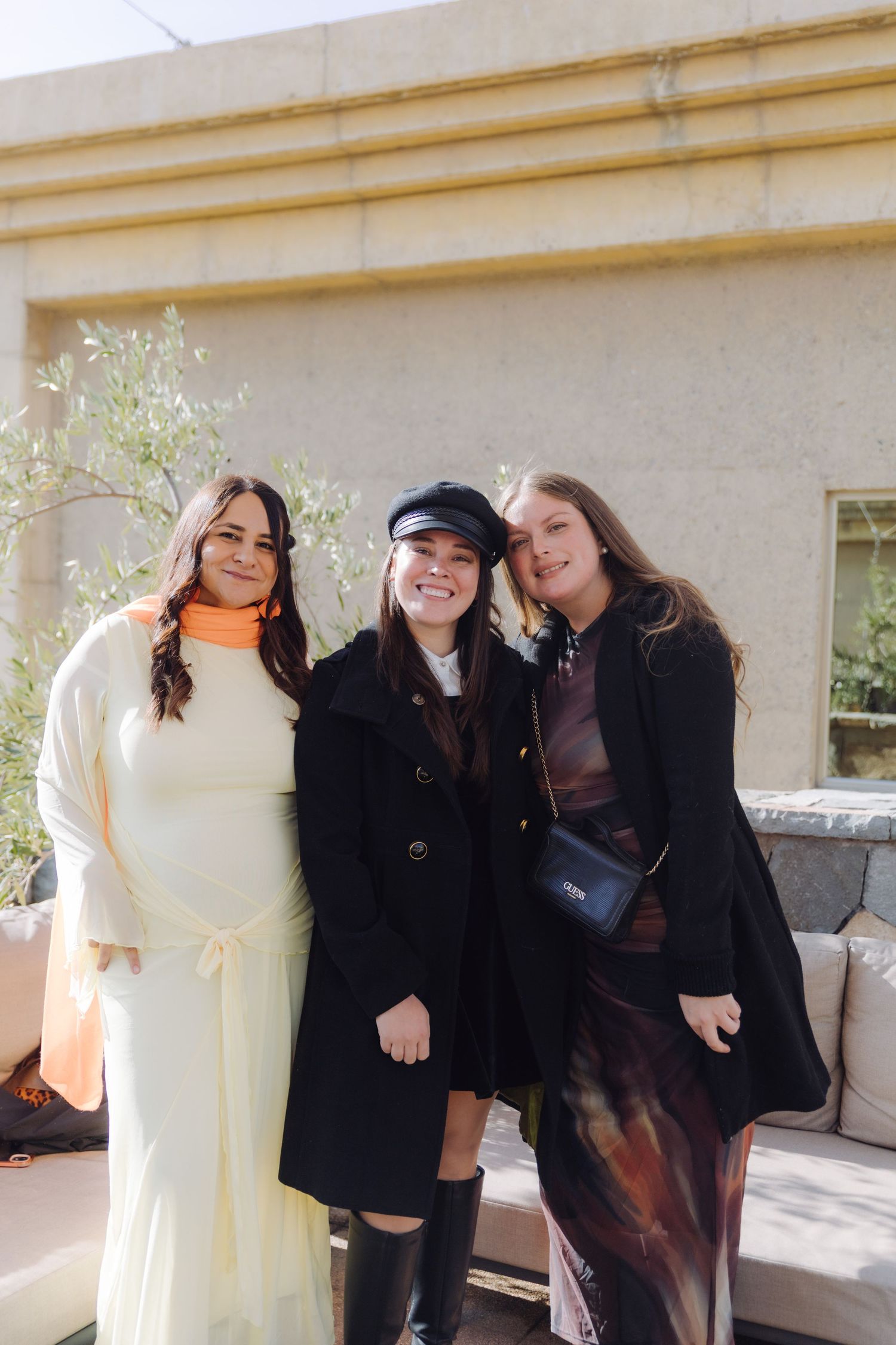 Three young women in elegant outfits smiling together at outdoor event venue.