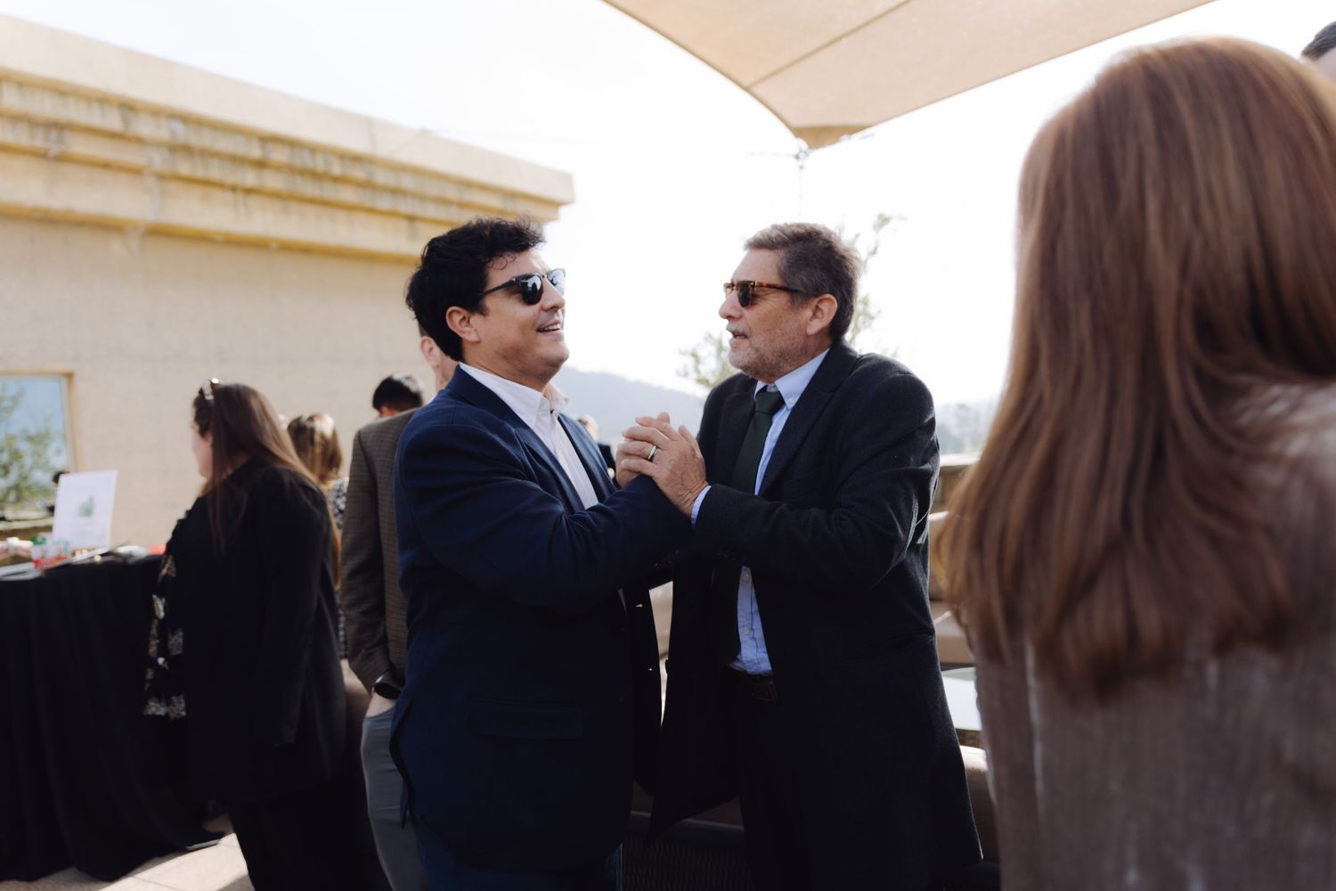 Two men in suits wearing sunglasses conversing at an outdoor formal event.