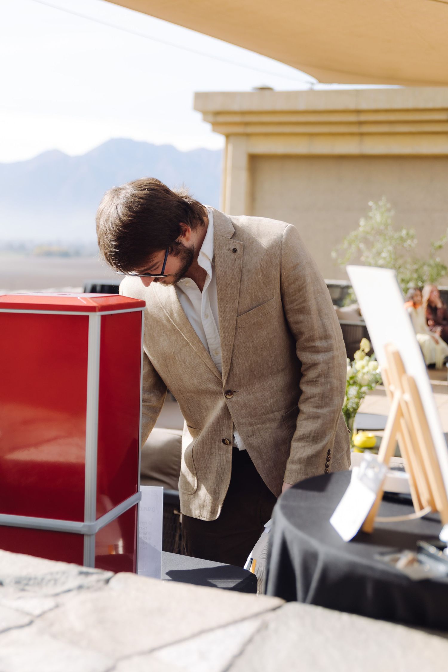 Man in beige blazer leaning over red display box at outdoor event table.