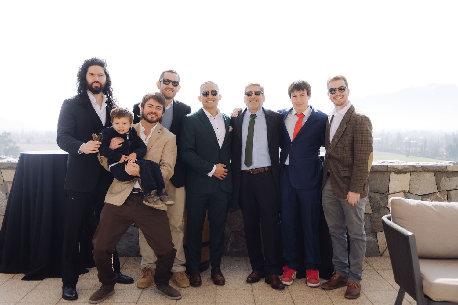 Group of eight men in formal attire posing together at outdoor rooftop gathering.