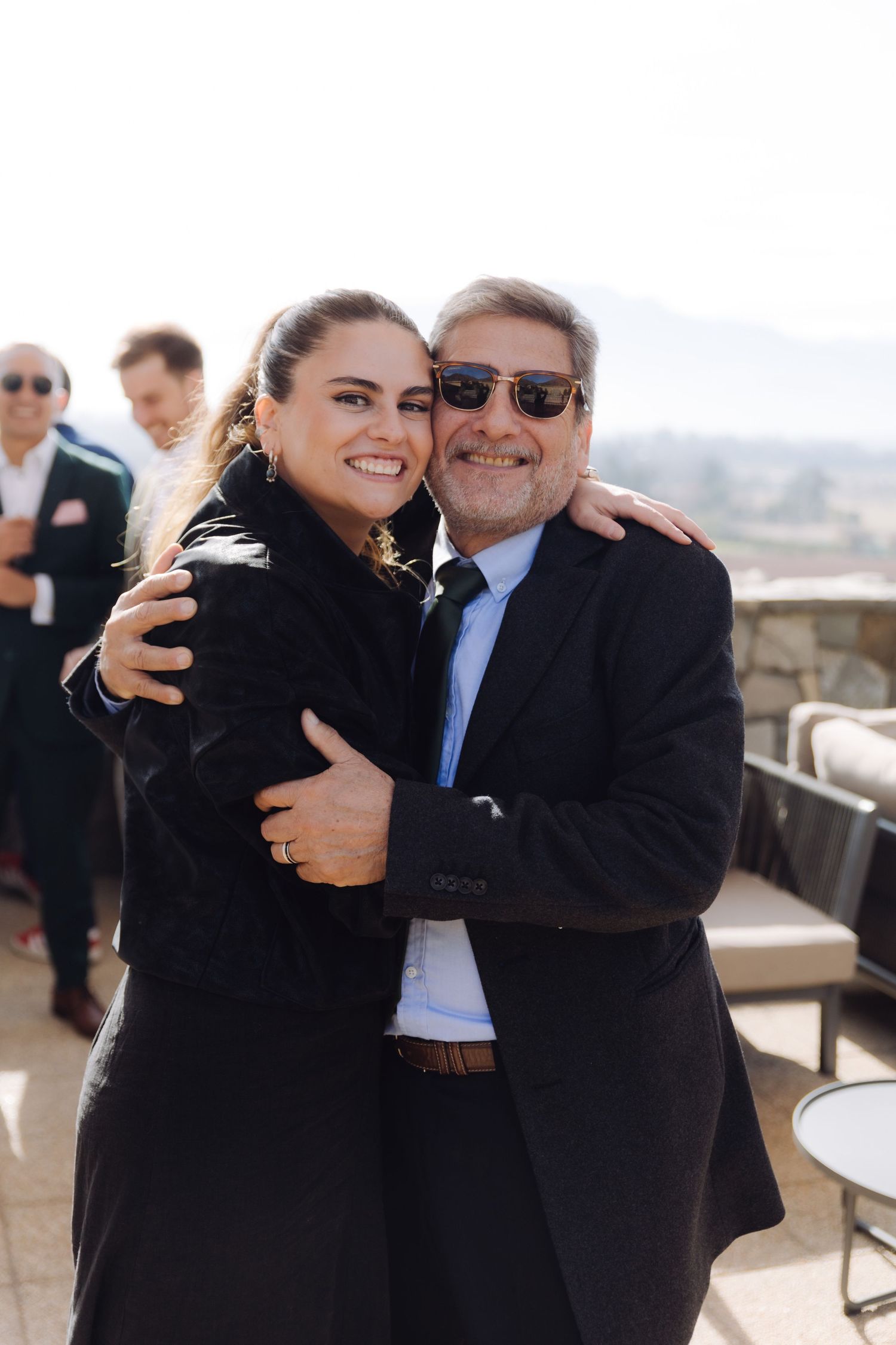 Smiling woman and older man in black coats embracing warmly at outdoor event.