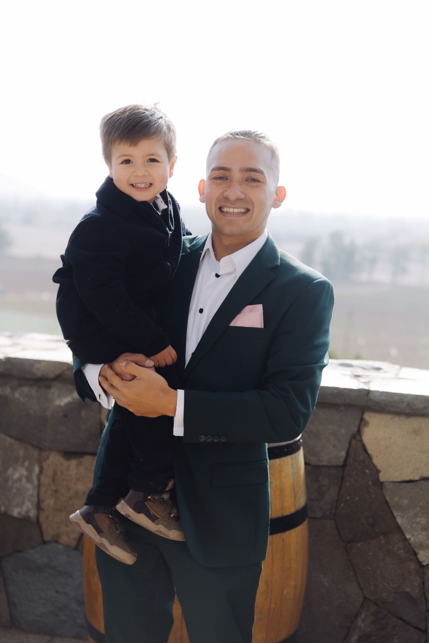 Smiling man in green suit holds young boy in black outfit at outdoor stone venue.
