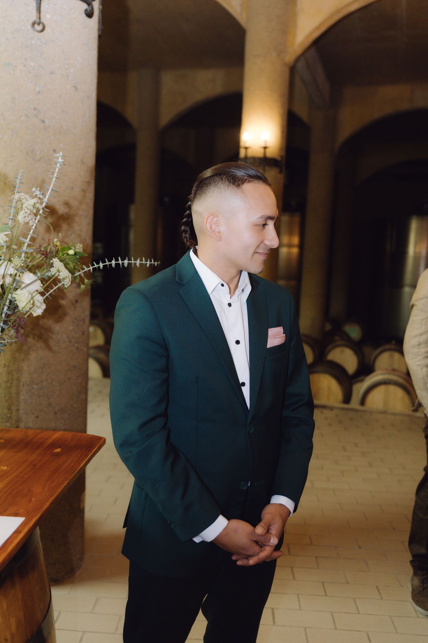 Groom in green suit with pink pocket square stands in rustic winery ceremony venue.