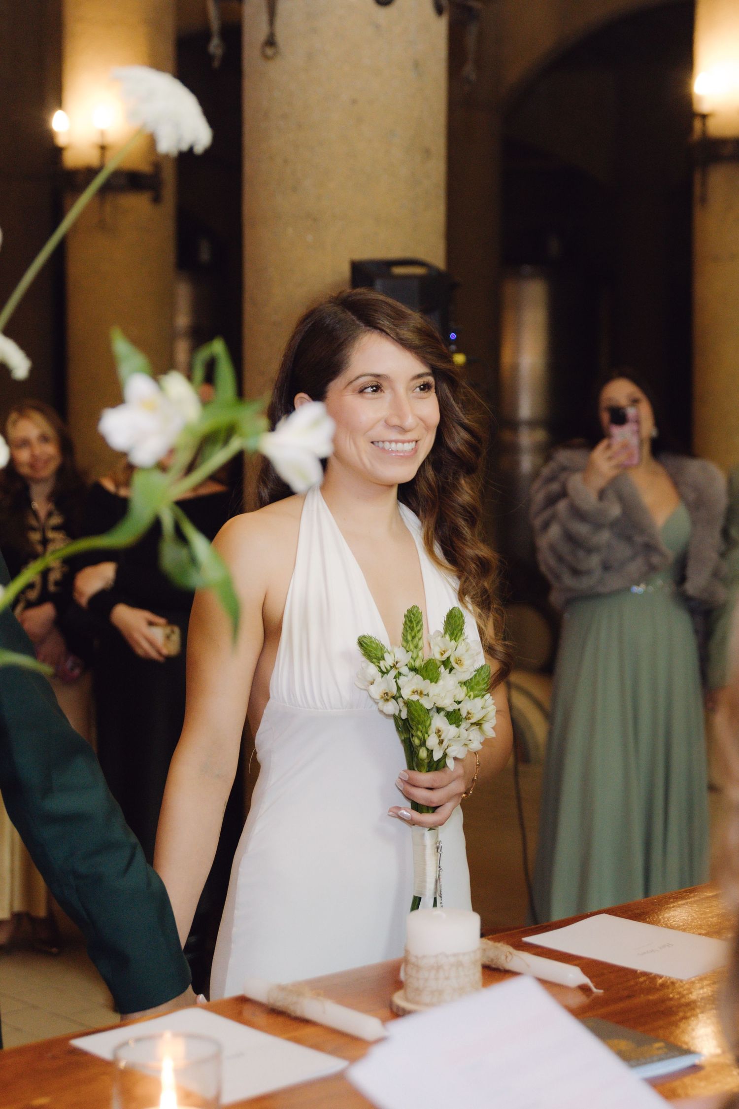 Smiling bride in white halter dress holds bouquet at winery wedding ceremony.