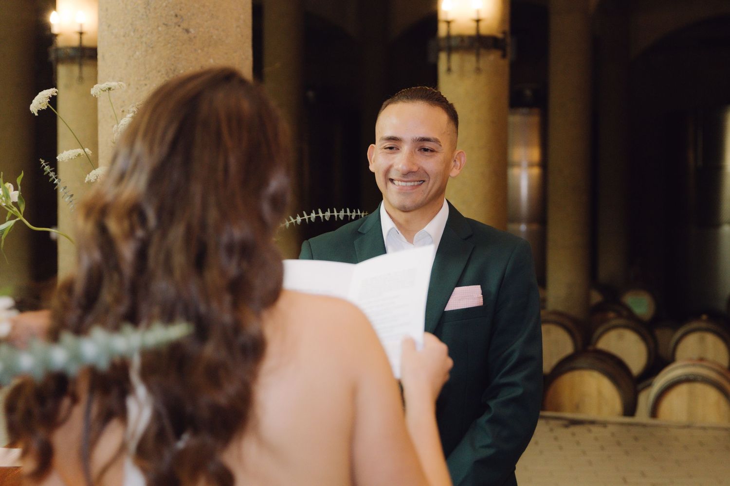 Groom in green suit smiles joyfully at bride during winery wedding ceremony.