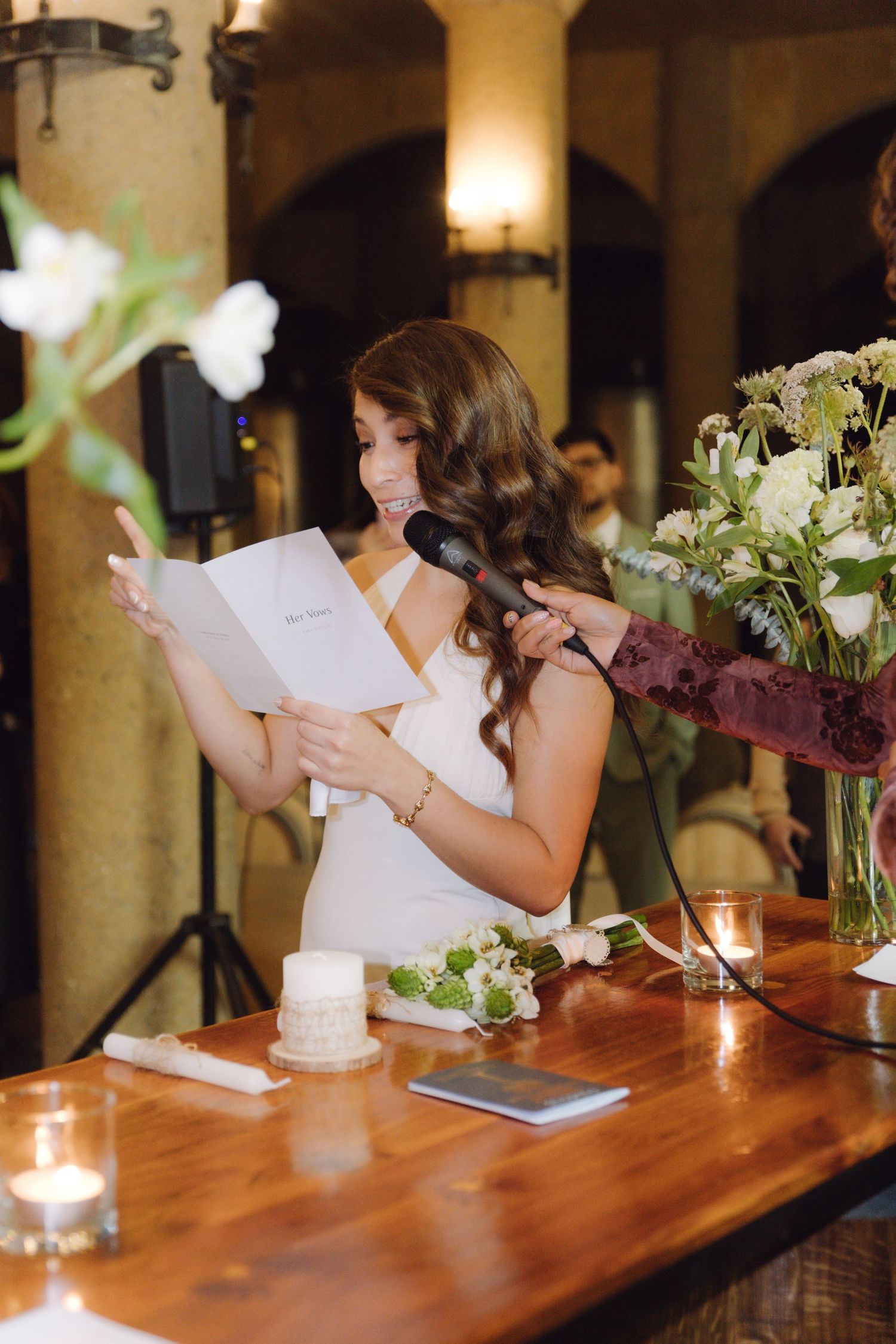 Bridesmaid delivers heartfelt speech at wedding reception holding microphone and notes at floral decorated wooden podium.