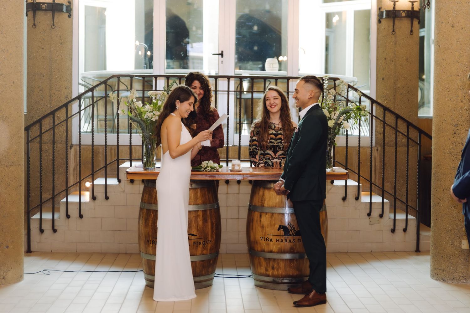 Bride and groom exchange vows at rustic wedding ceremony with wooden barrel altar and elegant staircase backdrop.