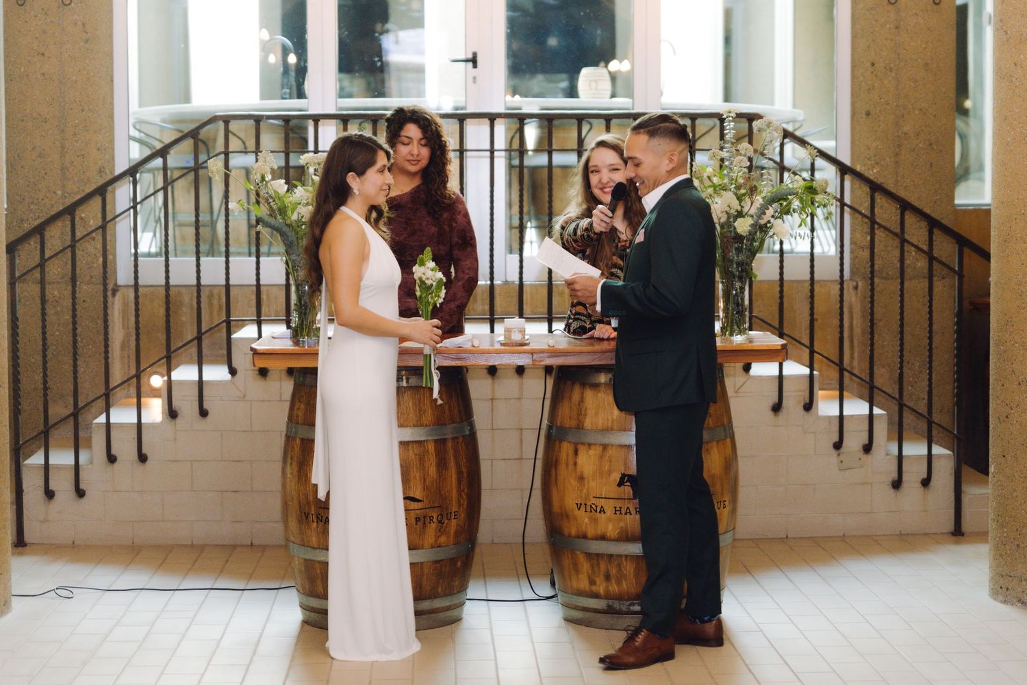 Couple exchanges rings during intimate wedding ceremony at rustic venue with wooden barrels and wrought iron staircase.
