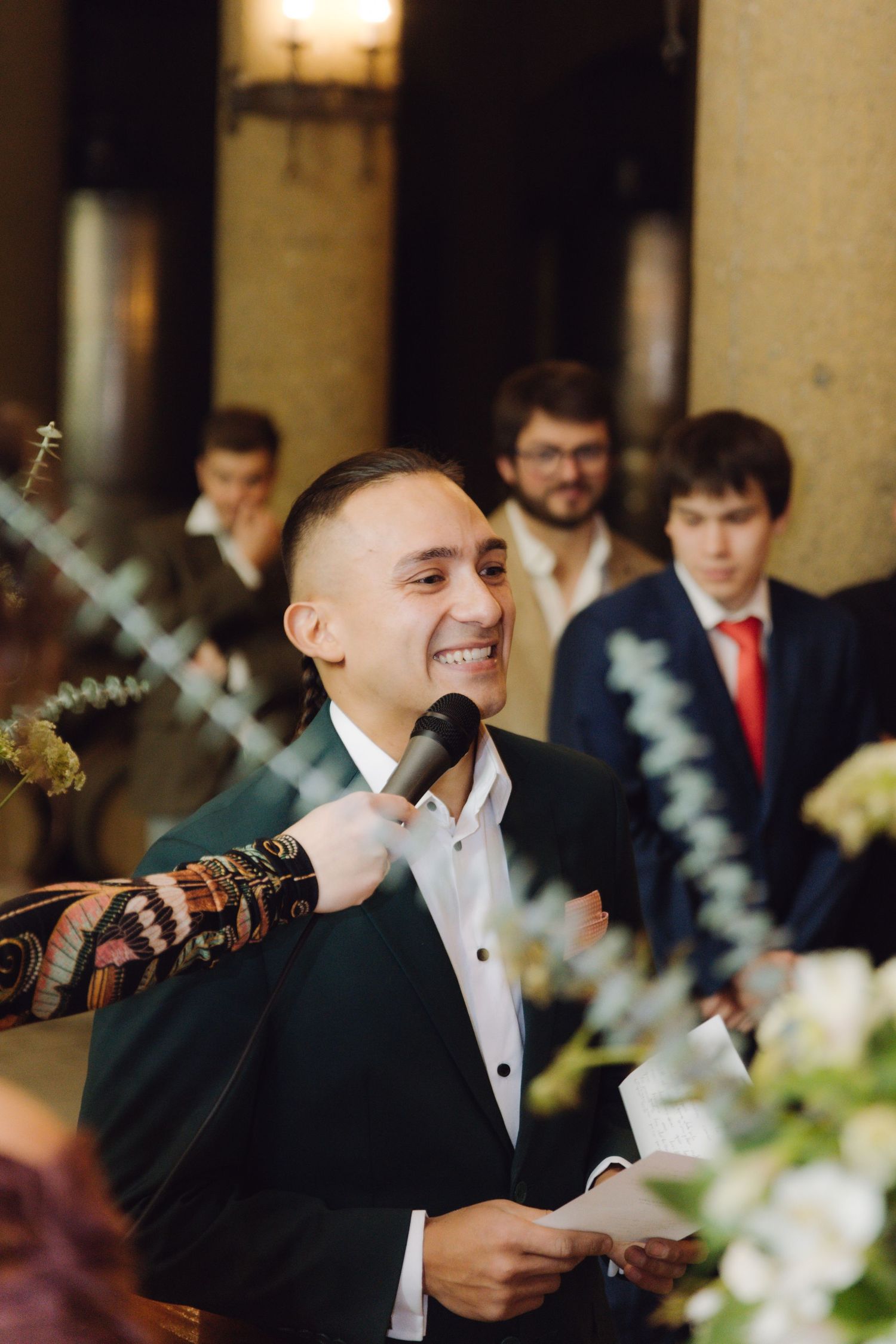 Smiling groomsman delivers wedding speech into microphone holding paper notes at elegant indoor reception venue.