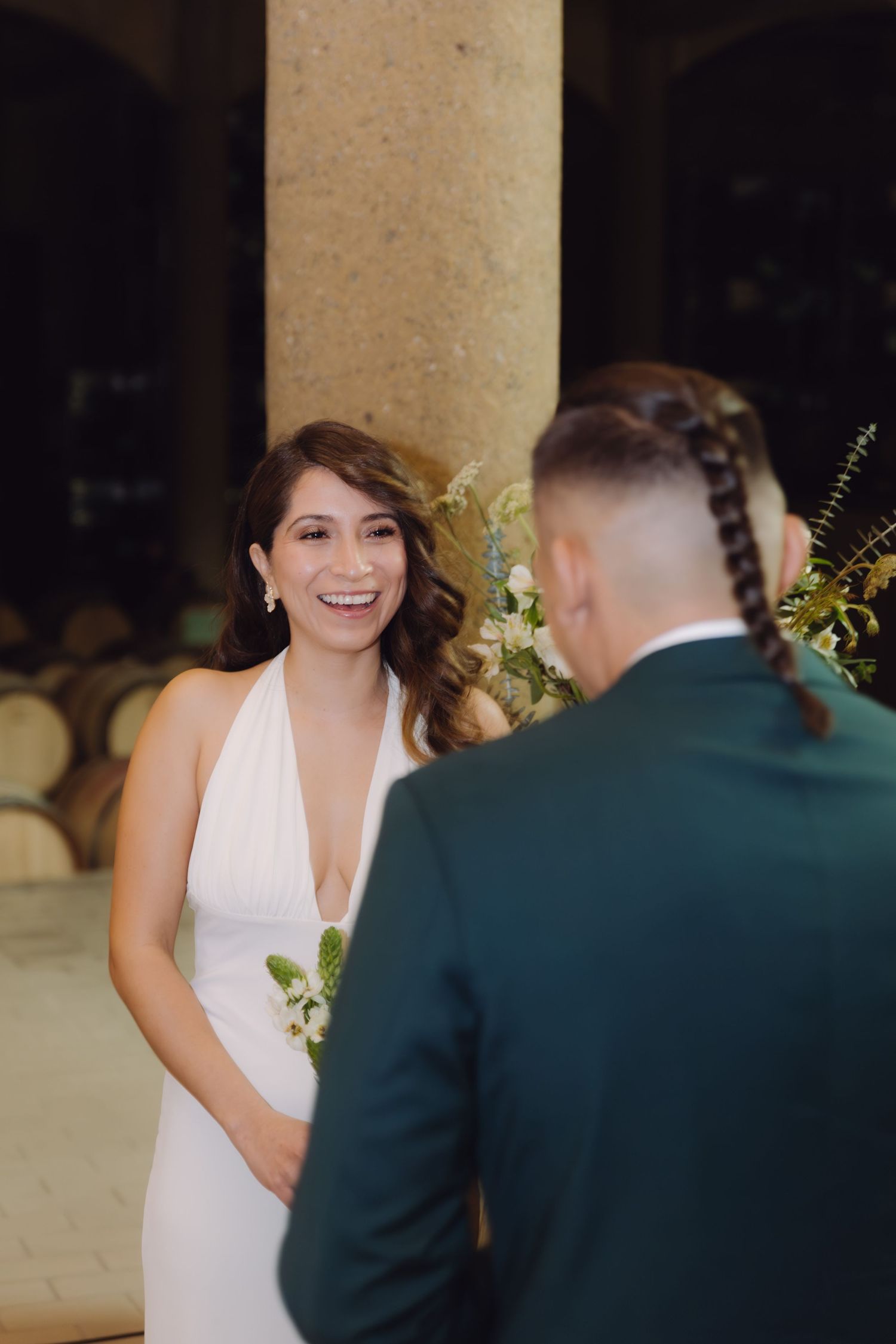 Radiant bride in white halter gown smiles joyfully at groom during wedding ceremony in elegant venue.