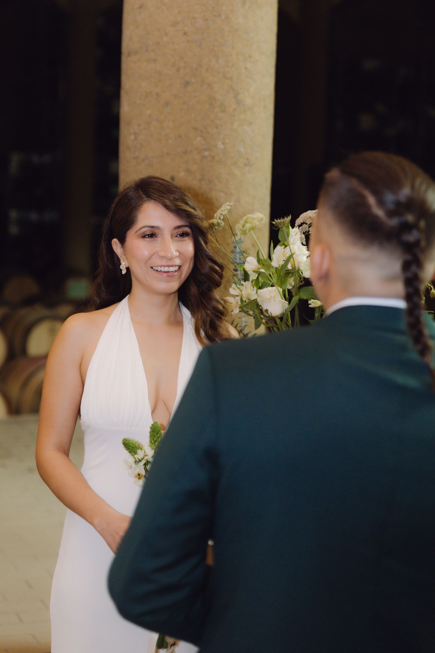 Beautiful bride in white halter dress smiles warmly at groom during romantic wedding ceremony indoors.