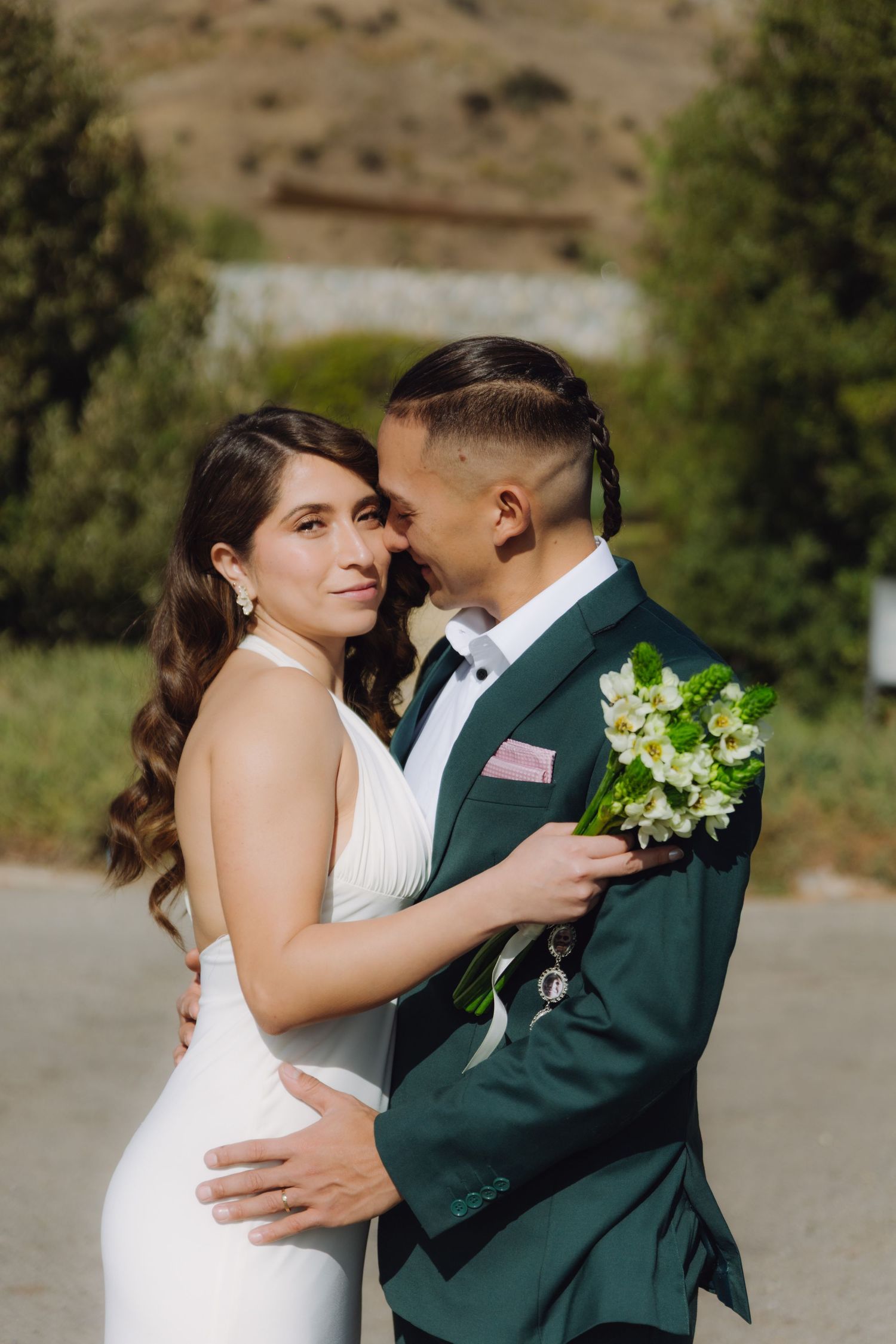 Bride and groom share an intimate moment outdoors, she in a white halter gown, he in a green suit holding a bouquet.