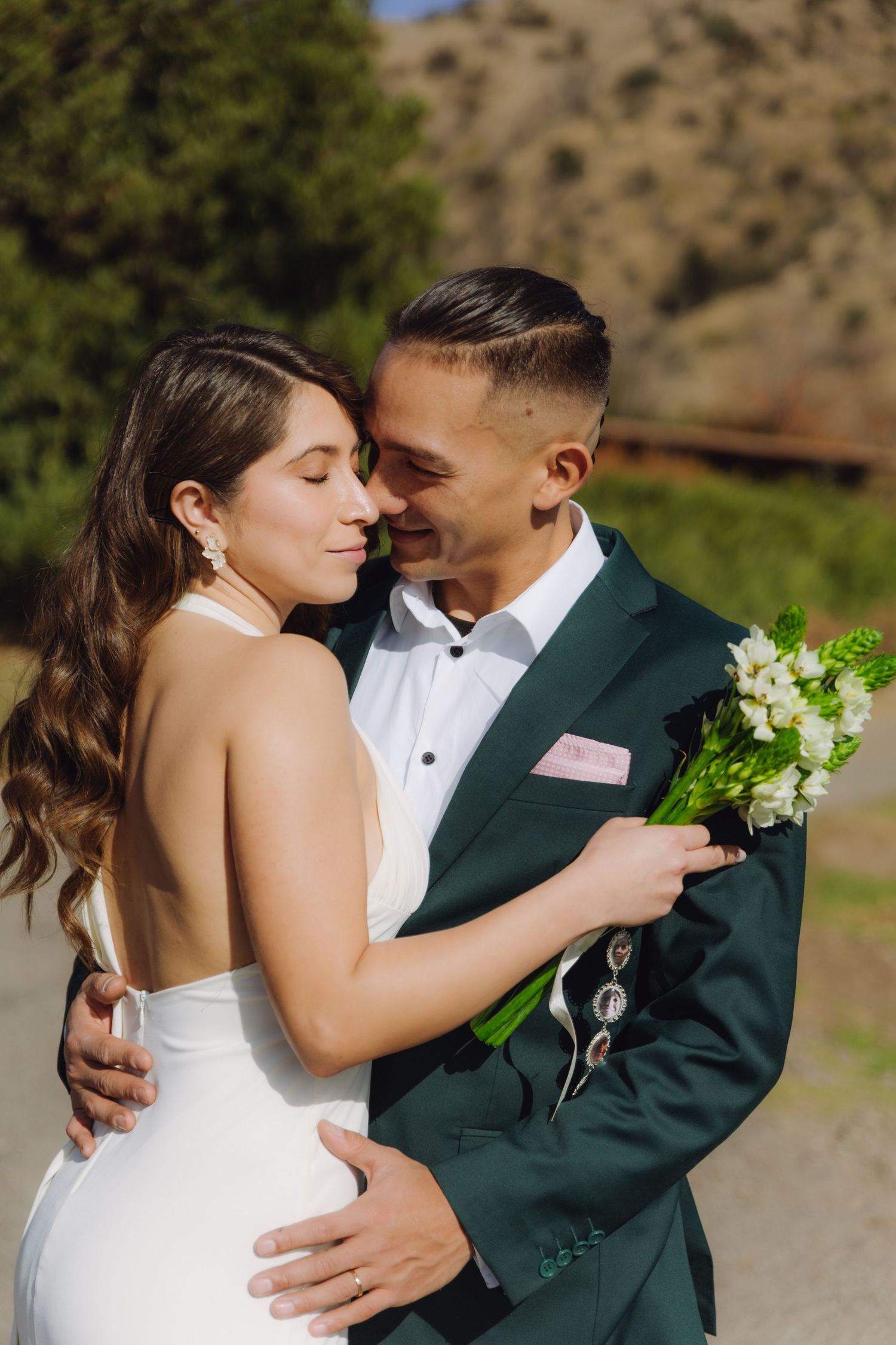 Wedding couple embracing outdoors, bride in backless white dress and groom in green suit holding white floral bouquet.