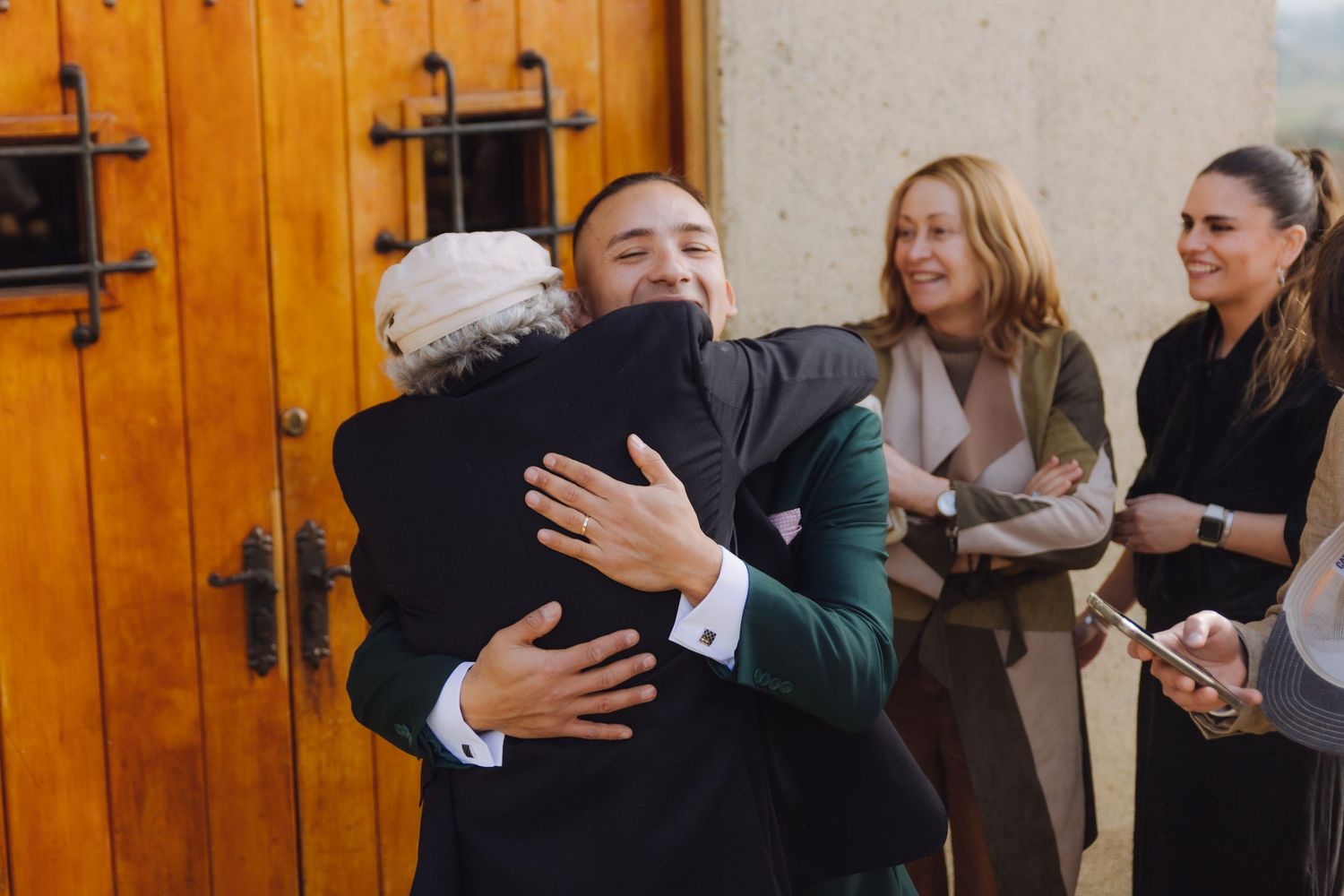 Groom in green suit shares a warm hug with a guest outside wooden doors at a wedding celebration.