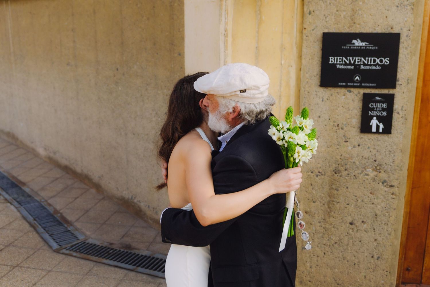 Bride in white dress embraces an older guest holding white flowers outside a building with a Bienvenidos sign.