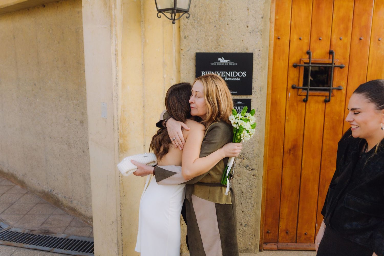 Bride in white dress shares a warm embrace with a female guest outside a venue entrance at her wedding.
