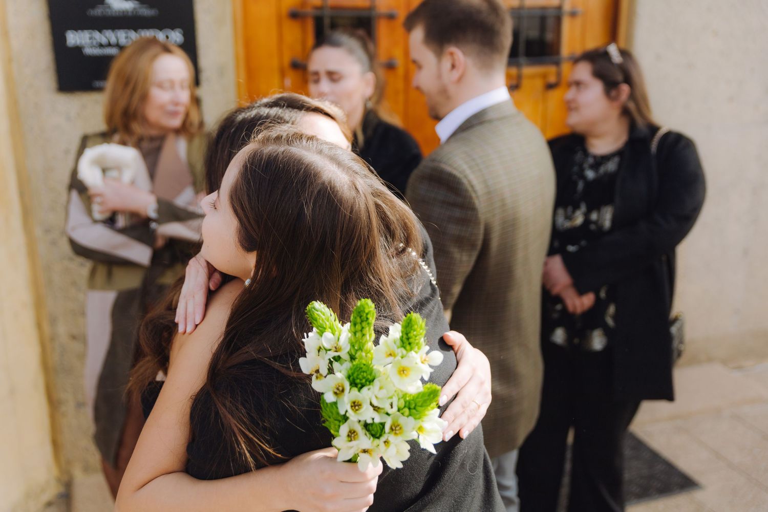 Bride holding white floral bouquet receives a heartfelt hug from a guest outside a wedding venue.