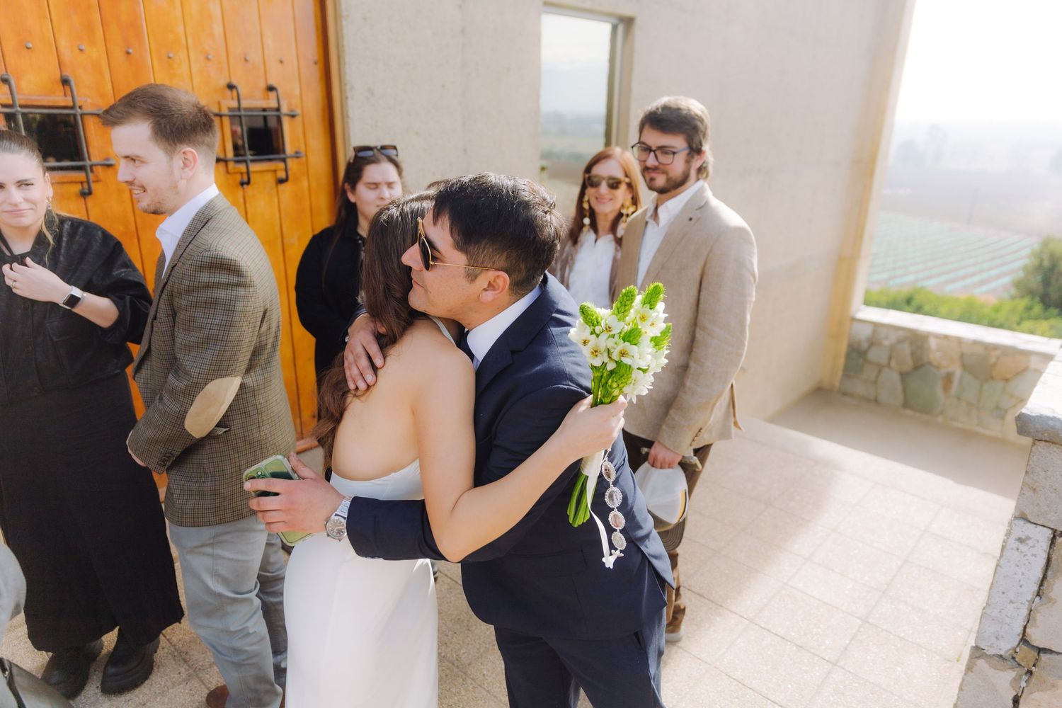 Bride and groom share a kiss outside a stone venue entrance surrounded by wedding guests.