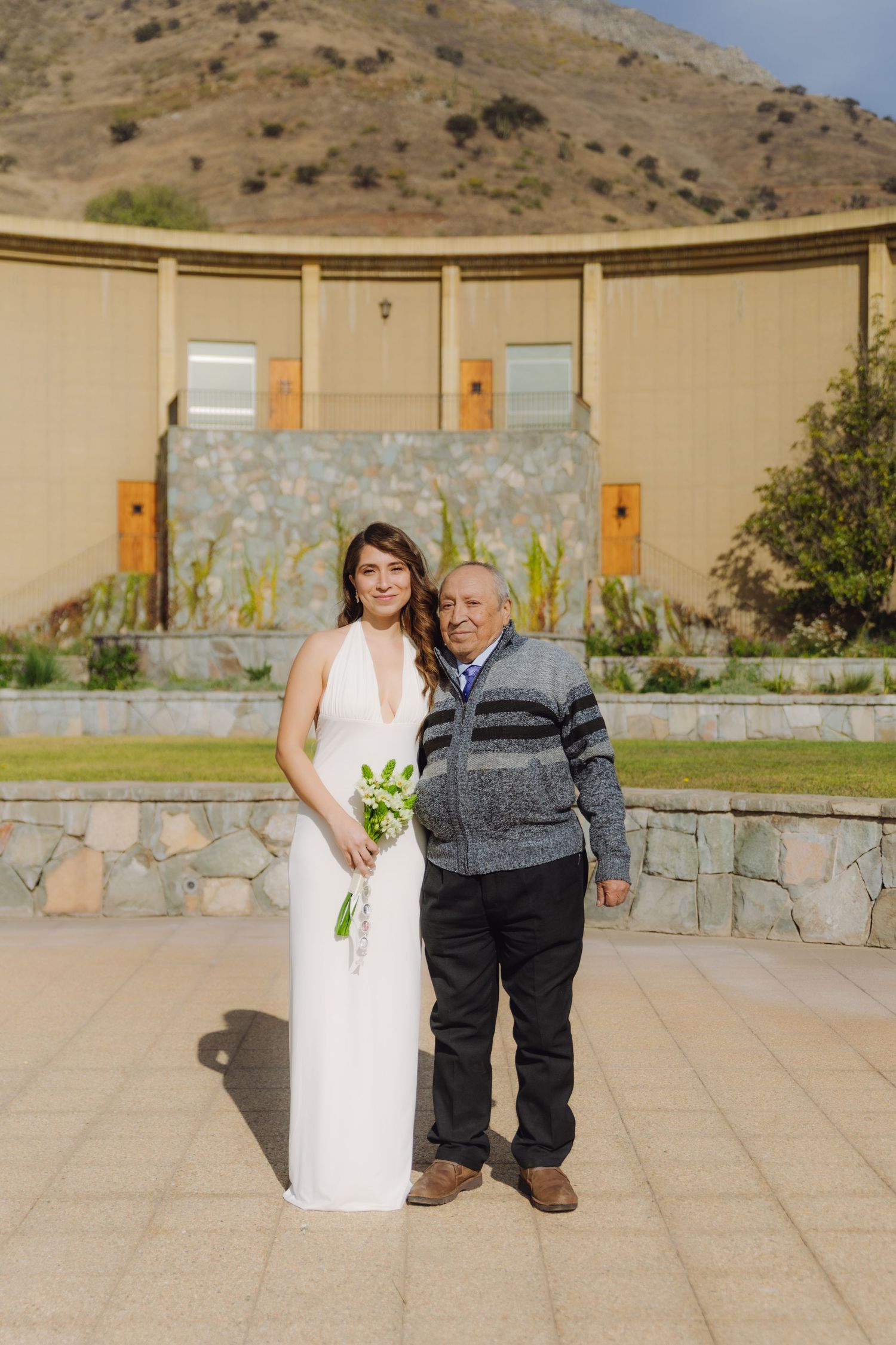 Bride in white halter gown holds green bouquet beside older man in gray sweater at outdoor stone venue.