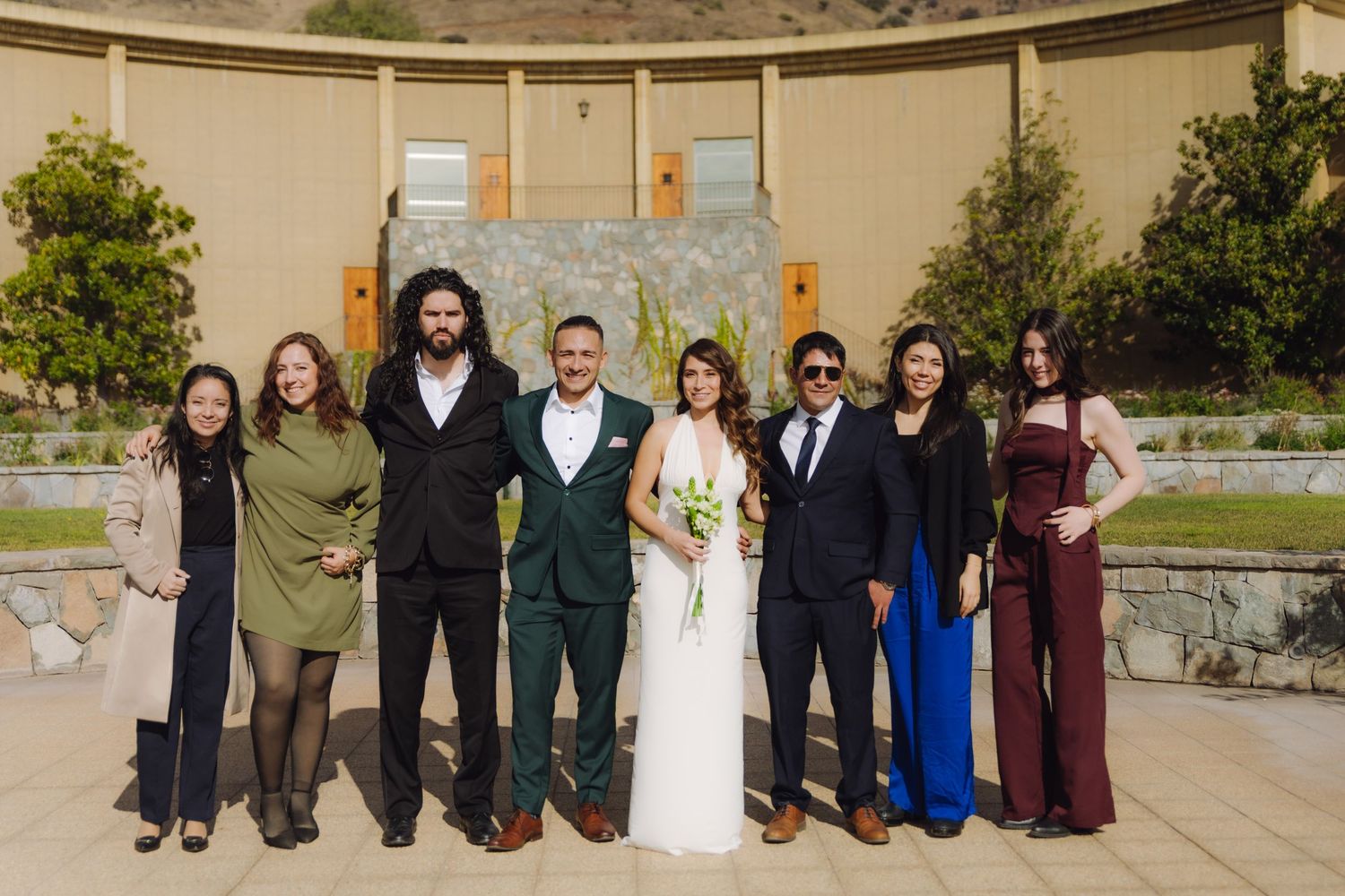 Wedding party of eight poses in colorful formal attire before stone fountain at outdoor mountain venue.