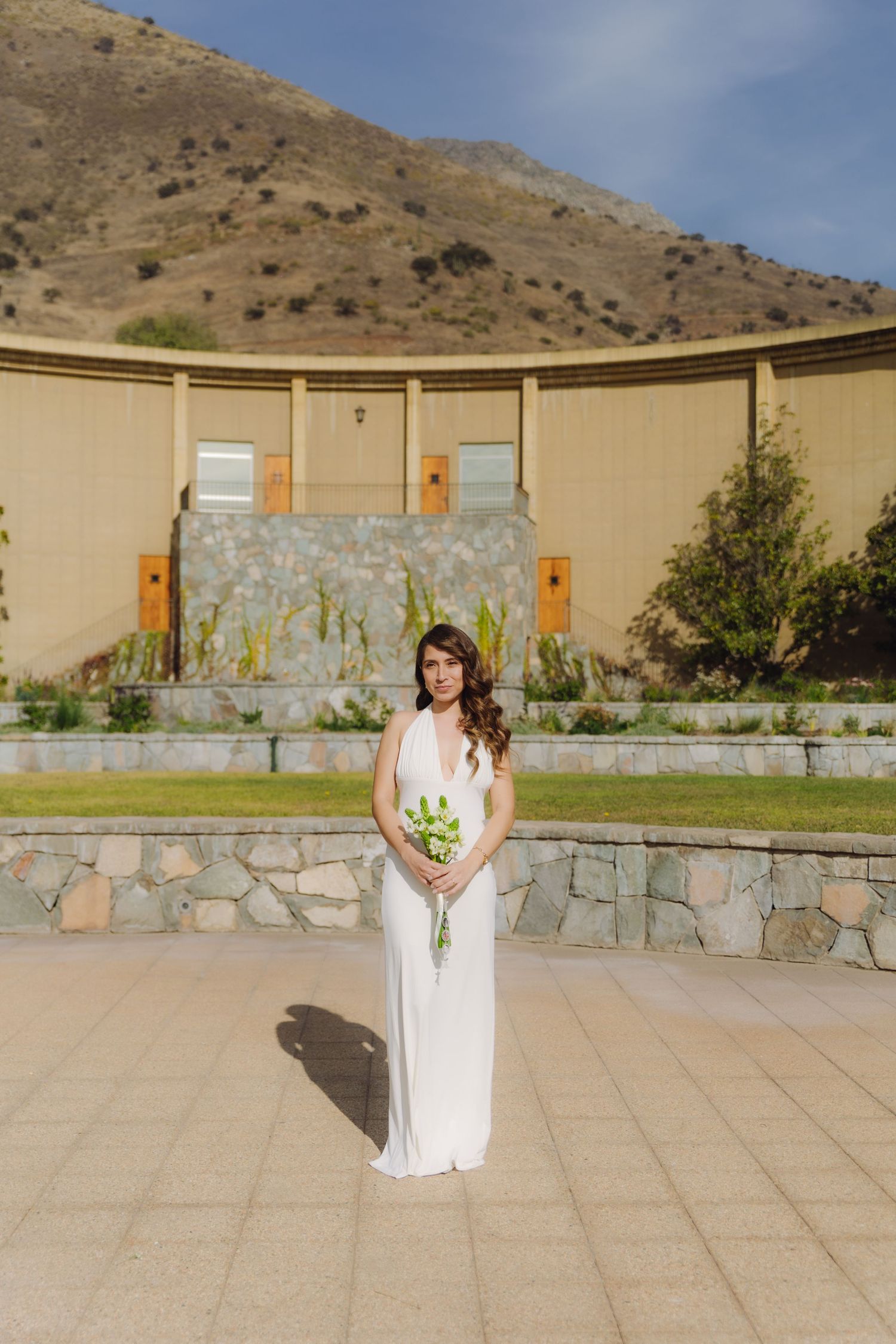 Elegant bride in sleeveless white gown holds green bouquet standing before stone wall with mountain backdrop.