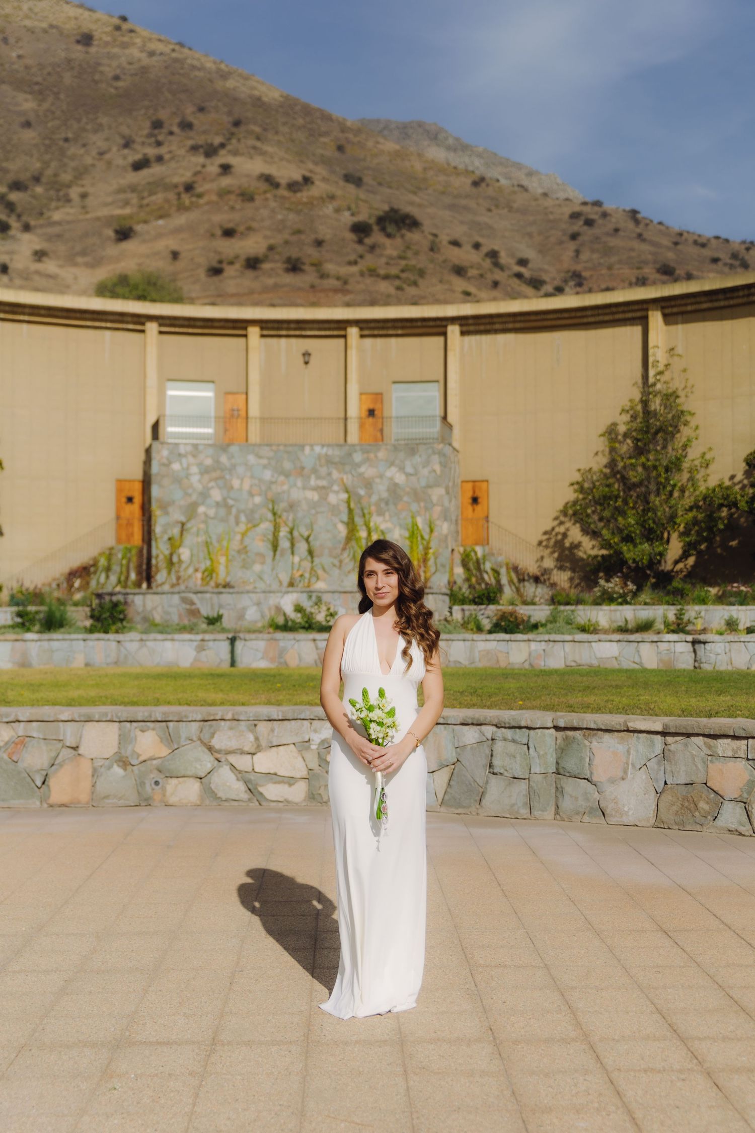 Bride in white halter dress holds green floral bouquet standing on paved plaza with mountain behind.