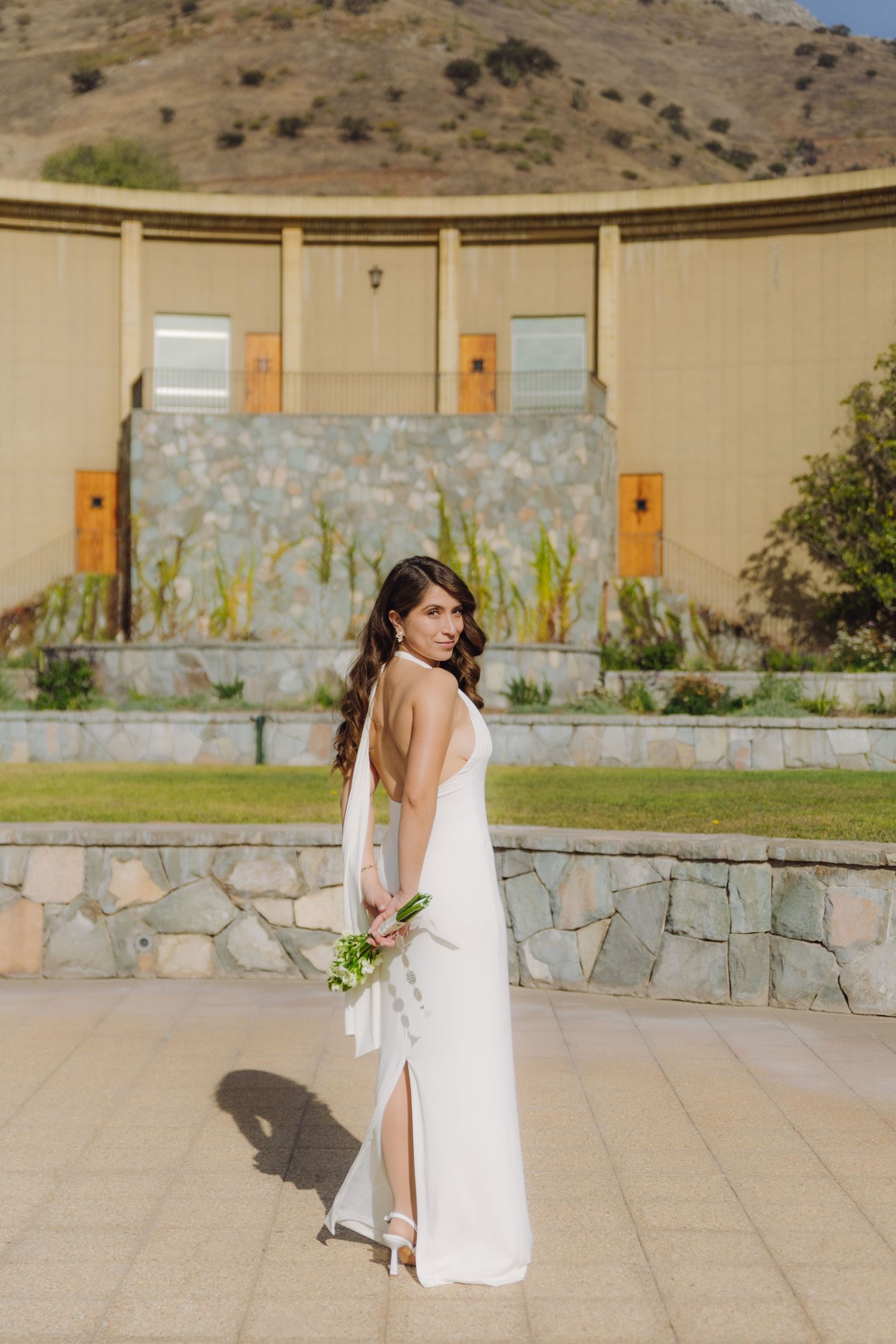 Bride in backless white gown glances over shoulder holding bouquet on sunny plaza with mountain view.