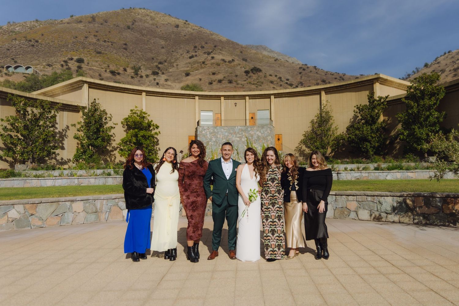 Wedding group of seven poses in formal attire outside stone chapel with mountain landscape background.