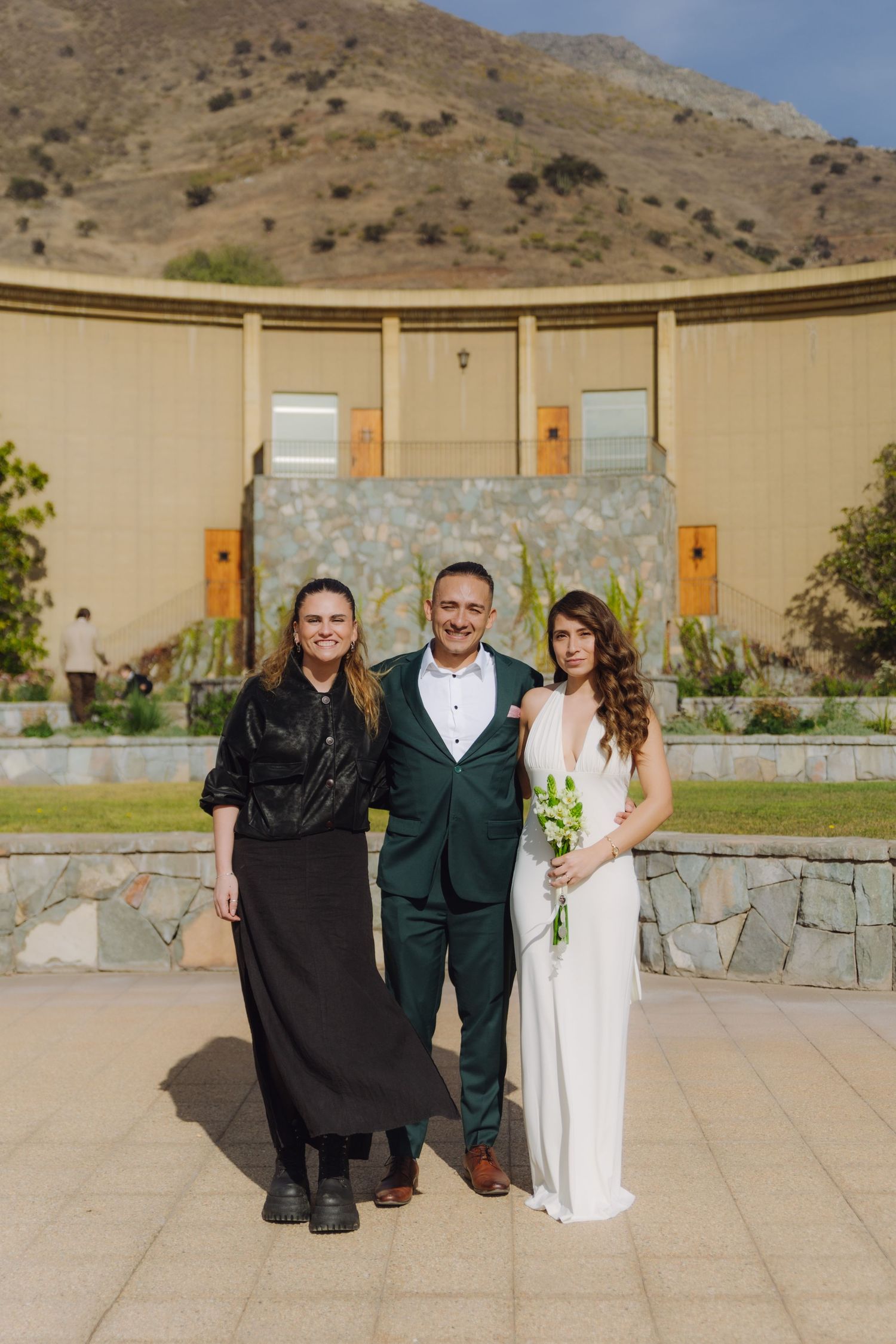Bride in white gown poses with groom in green suit and woman in black outfit at outdoor venue.