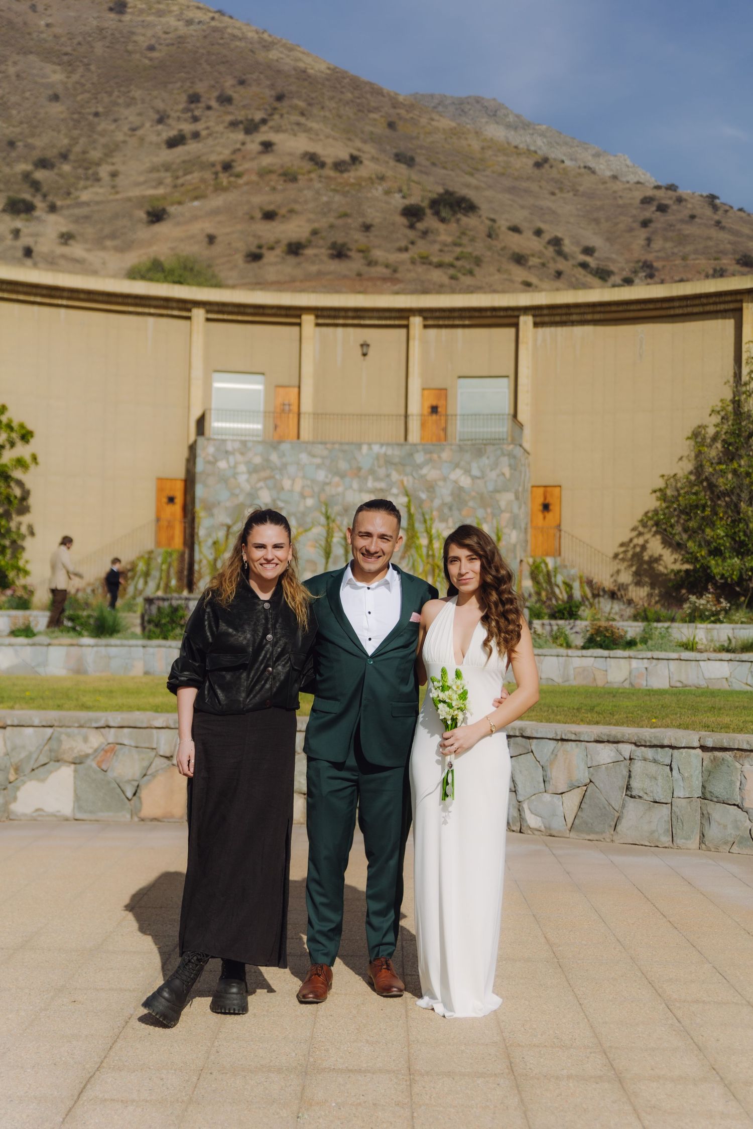 Smiling trio including bride, groom in green tuxedo, and woman in black pose at scenic outdoor venue.