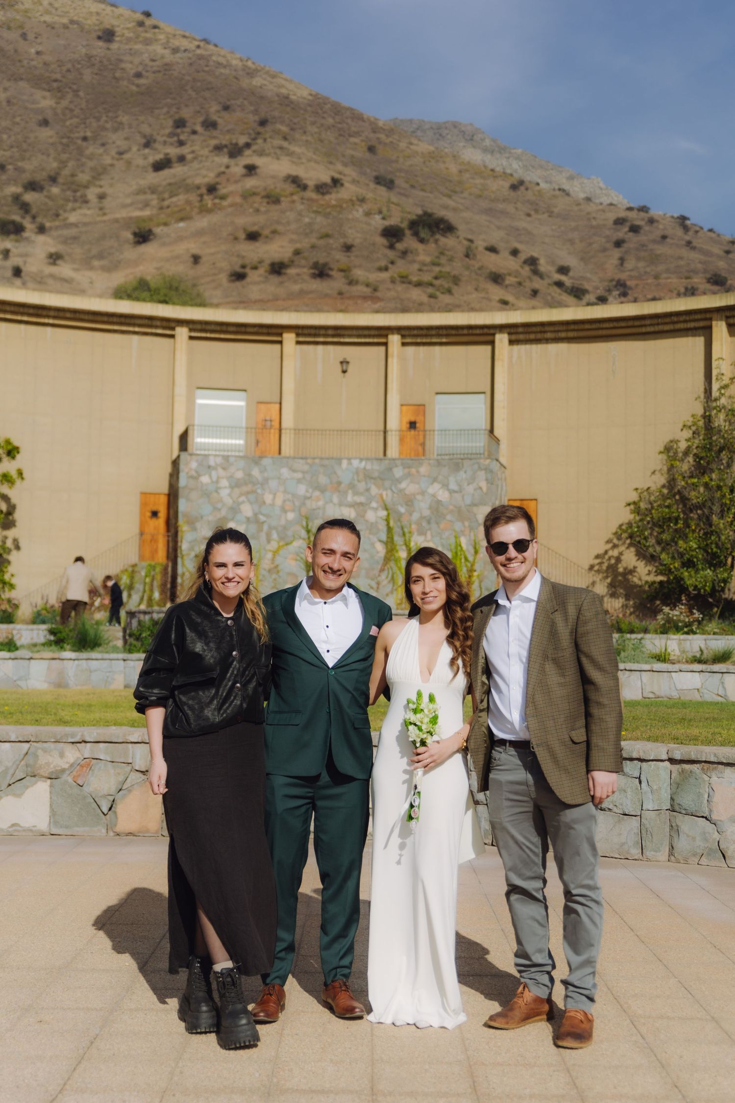 Wedding party of four poses outdoors at modern venue with mountain backdrop on sunny day.