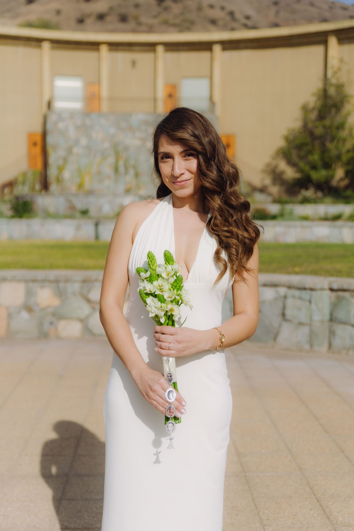 Smiling bride in elegant white halter wedding dress holds a delicate bouquet of white flowers outdoors.