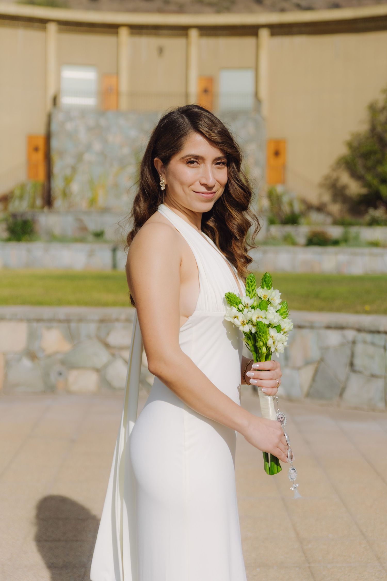 Bride in white halter gown with long train smiles over shoulder holding a white floral bouquet.