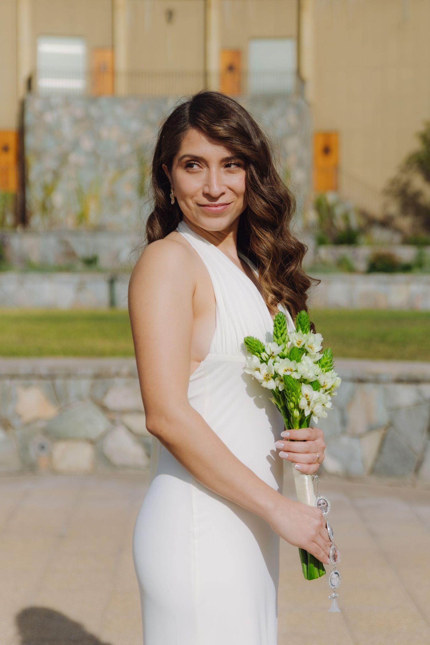 Bride in sleek white halter dress poses with a small white flower bouquet at an outdoor venue.