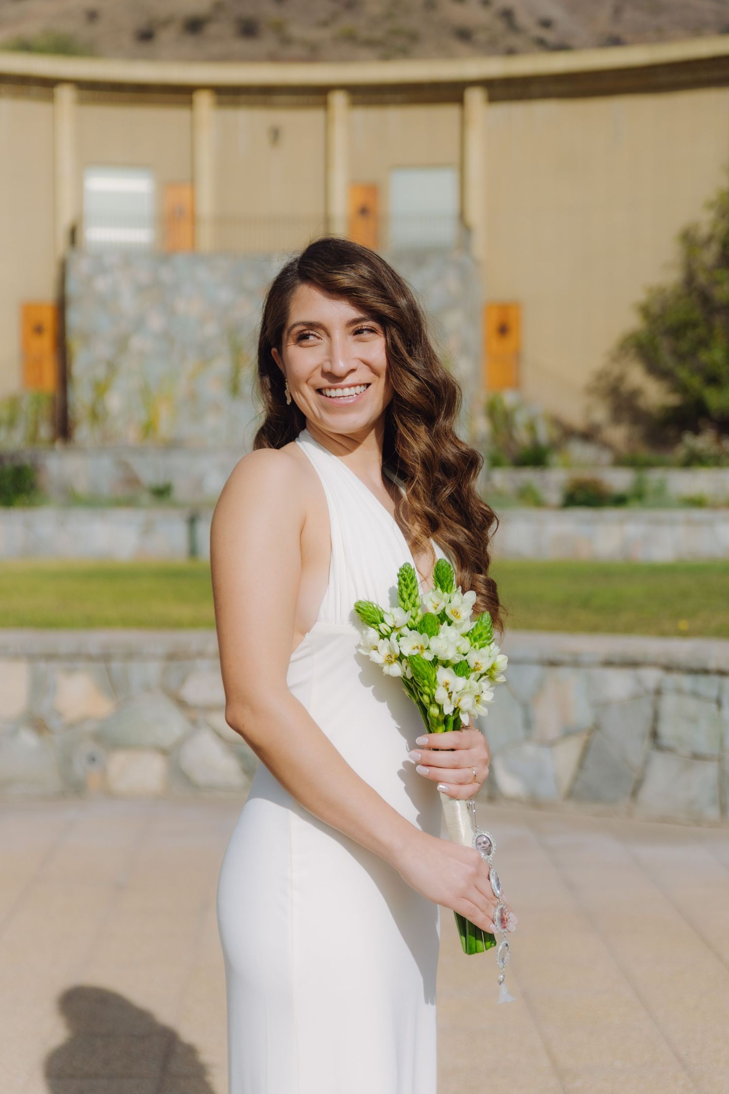 Radiant bride in white halter wedding dress smiles broadly while holding a white floral bouquet outdoors.
