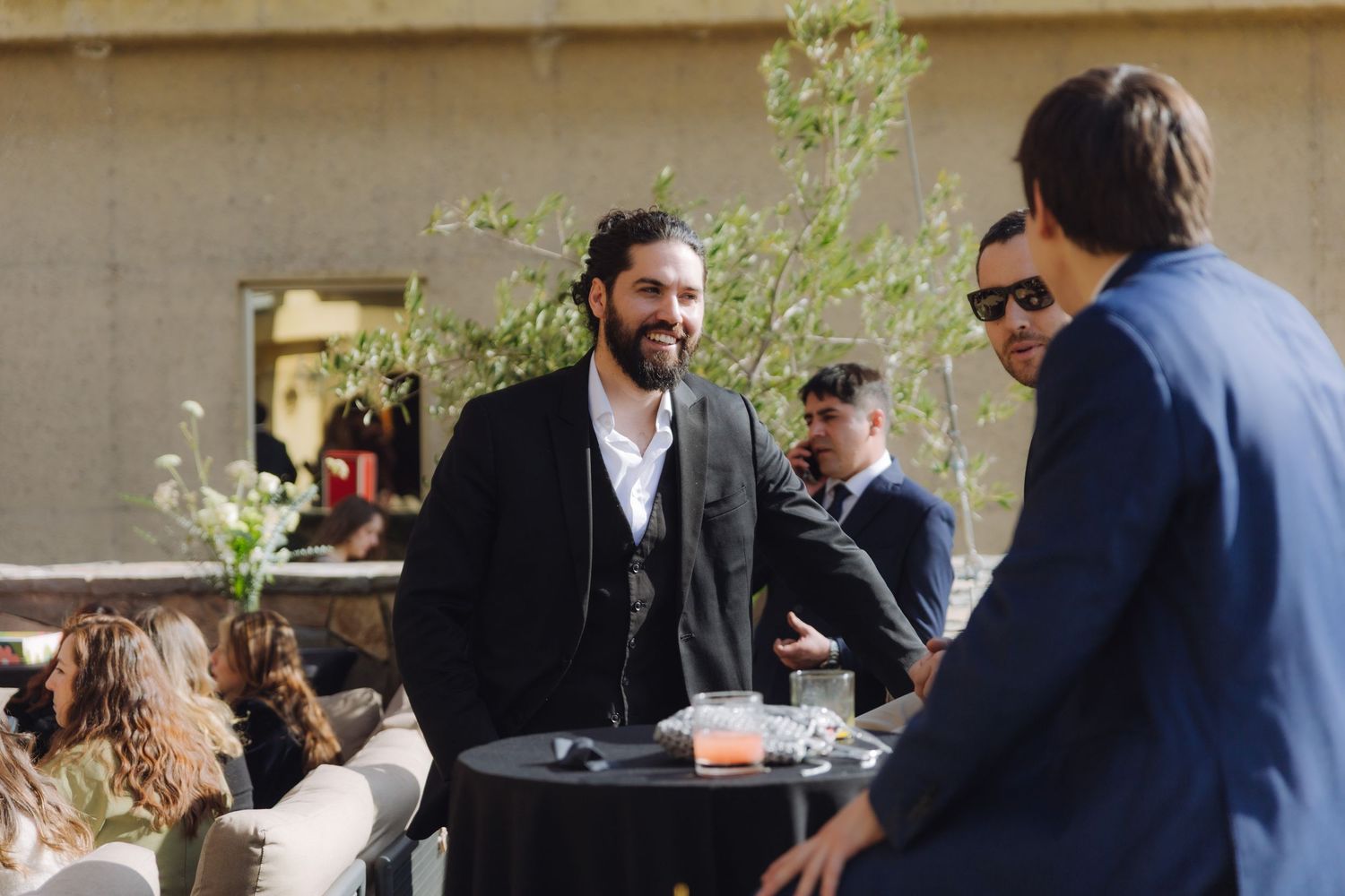 Bearded man in black suit smiles at guests during an outdoor wedding reception.