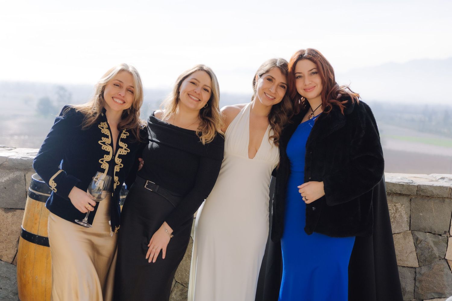 Four smiling women including a bride in white dress pose at an outdoor wedding venue.