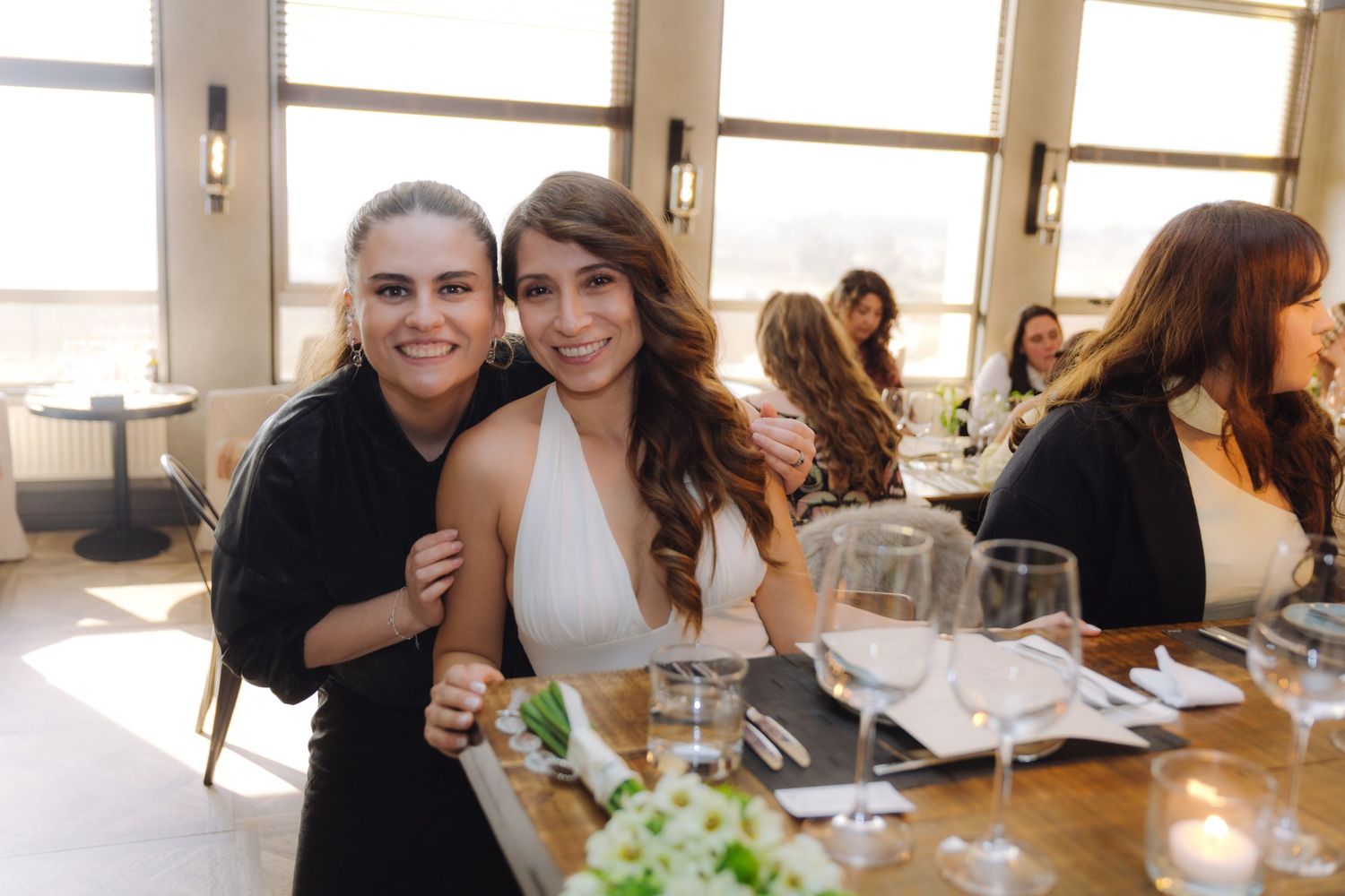Two women smile and pose together at an elegant restaurant dining table set with glassware.