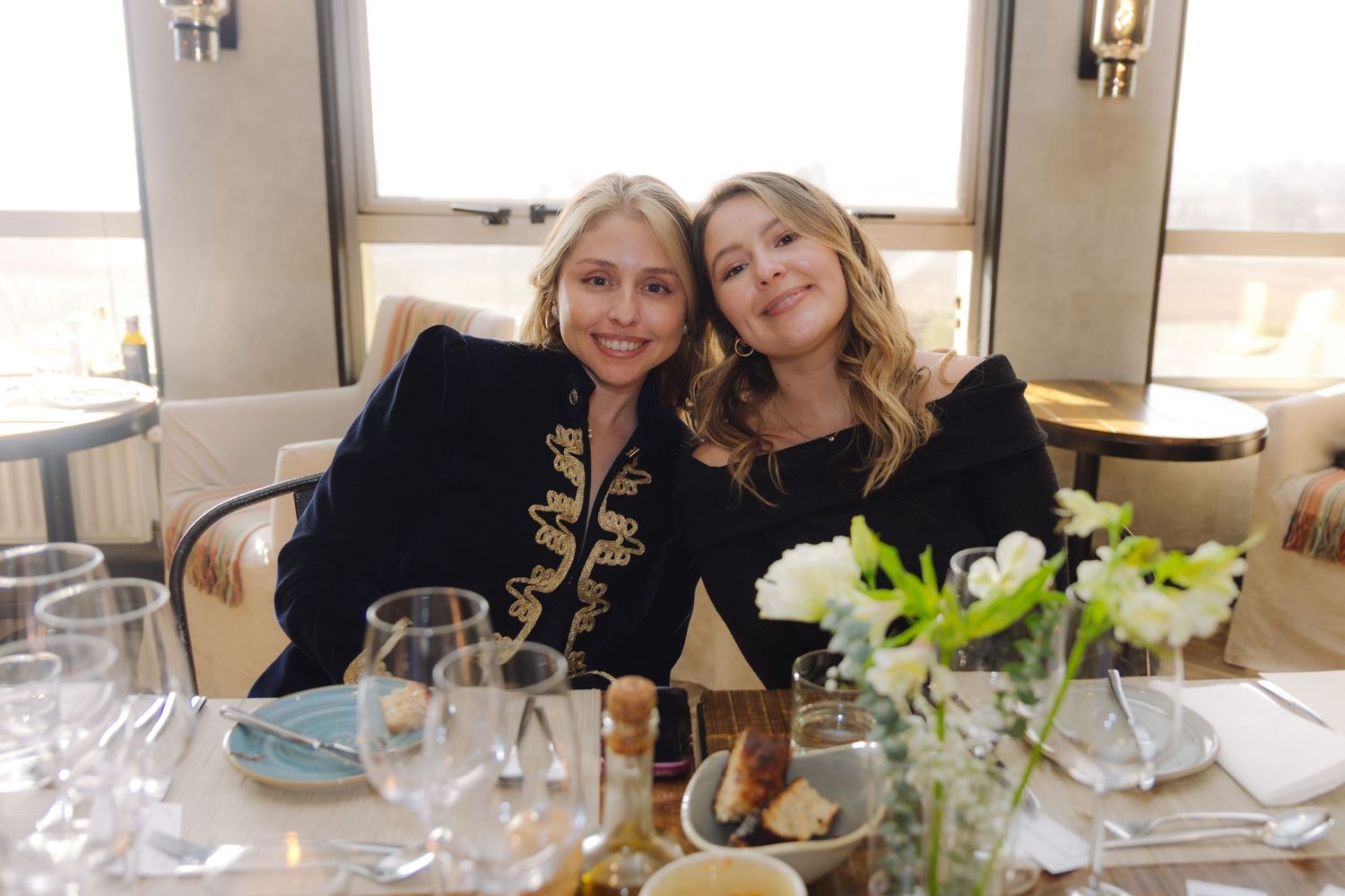 Two women in dark jackets smile together at a formal dining table with white flowers.