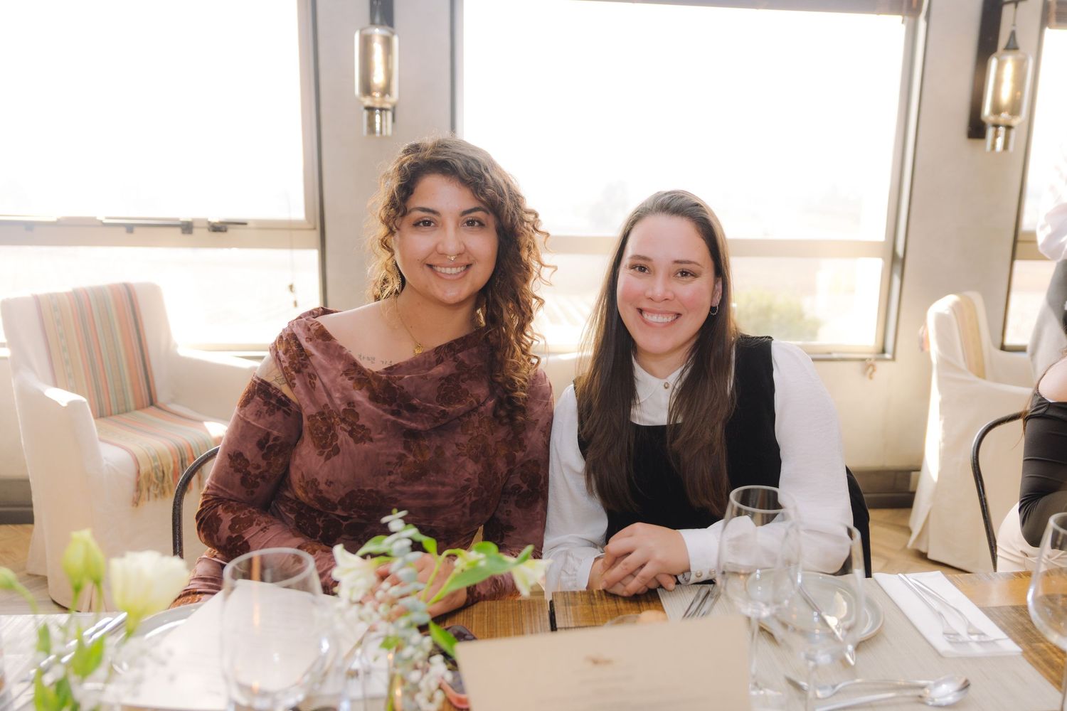 Two women smile at a restaurant table with elegant place settings and natural window light.