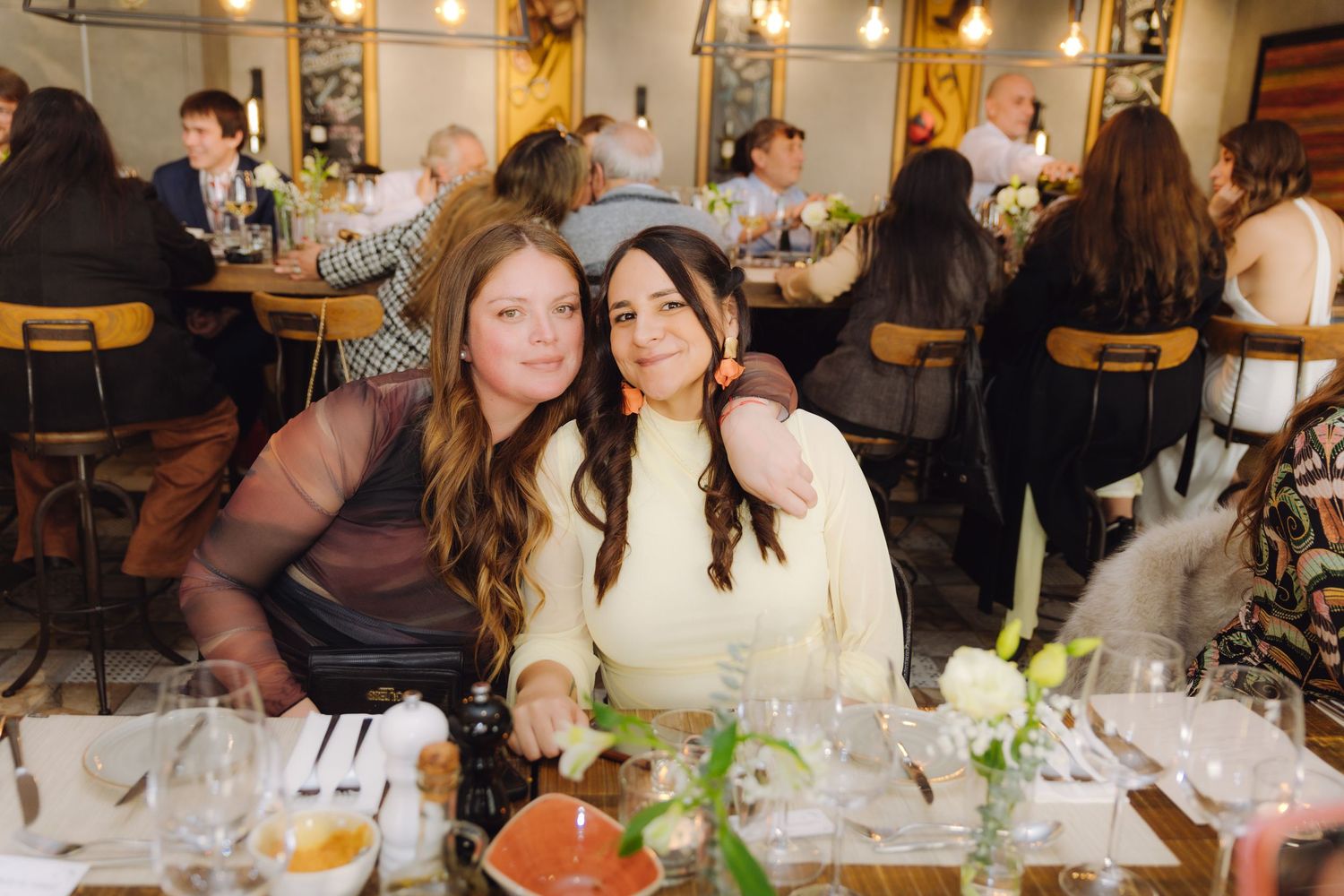 Two women smile together at a decorated dining table with white floral arrangements.
