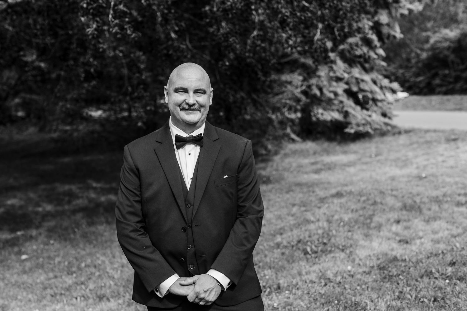 Man in tuxedo with bow tie smiling outdoors in black and white wedding portrait photo.
