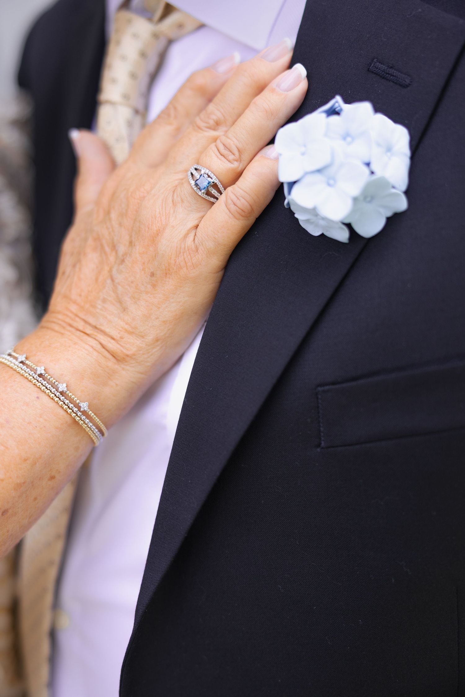 Close-up of bride's hand with diamond ring and bracelet resting on groom's navy suit with blue floral boutonniere.