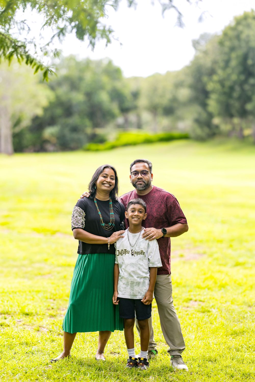 A family of three poses outdoors on green grass, with parents flanking their young son in a sunny park setting.