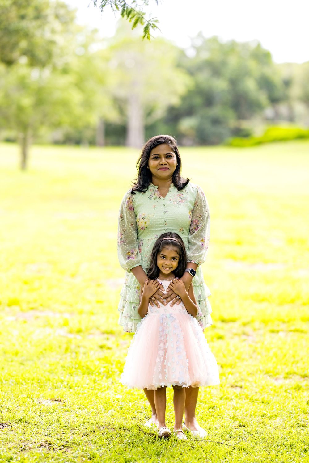 A woman in a floral kurta stands with a young girl in a pink dress on green grass in a park.